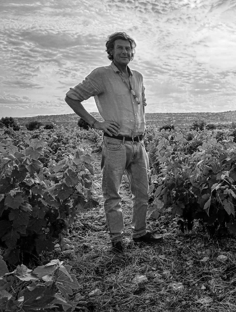 A man stands in a field of plants with a scenic sky in the background. He has curly hair, wears a button-up shirt and jeans, and has glasses hanging around his neck.