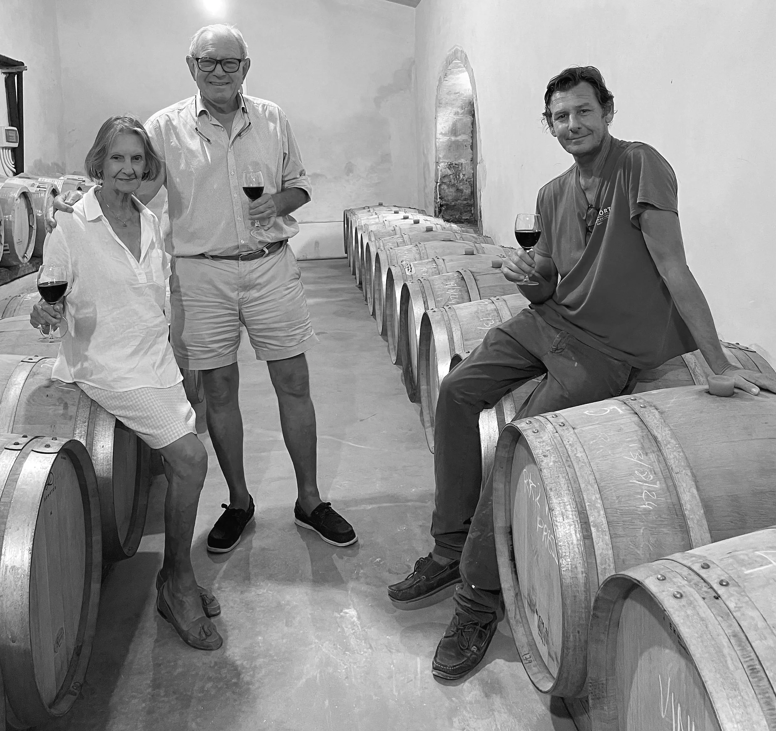 Four people standing in a wine cellar near barrels of wine, holding glasses of red wine, smiling.