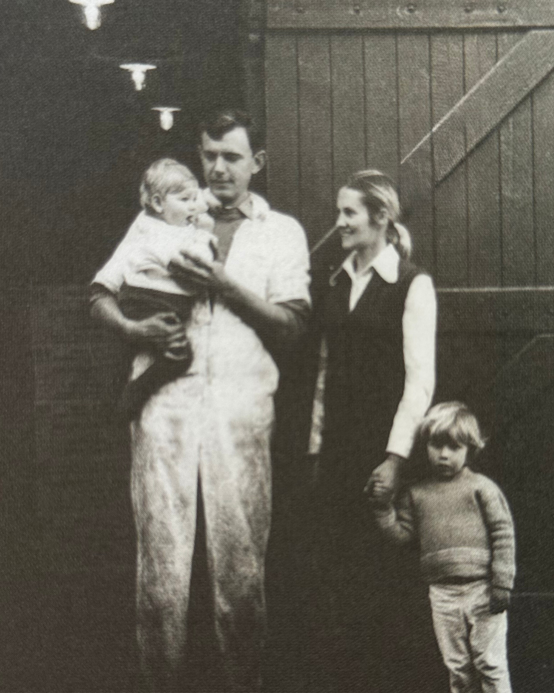 Black-and-white photo of three children and a woman outdoors. The woman is holding a young boy, and another girl is standing nearby. A wooden fence is visible in the background.