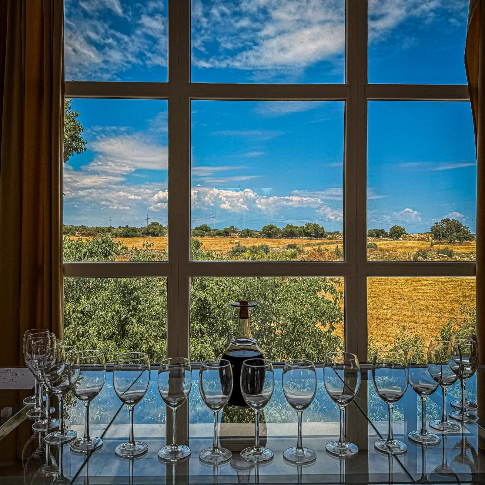 View through a large window showing a blue sky with scattered clouds, a field with trees and brush, and a row of wine glasses and a decanter on a table inside.