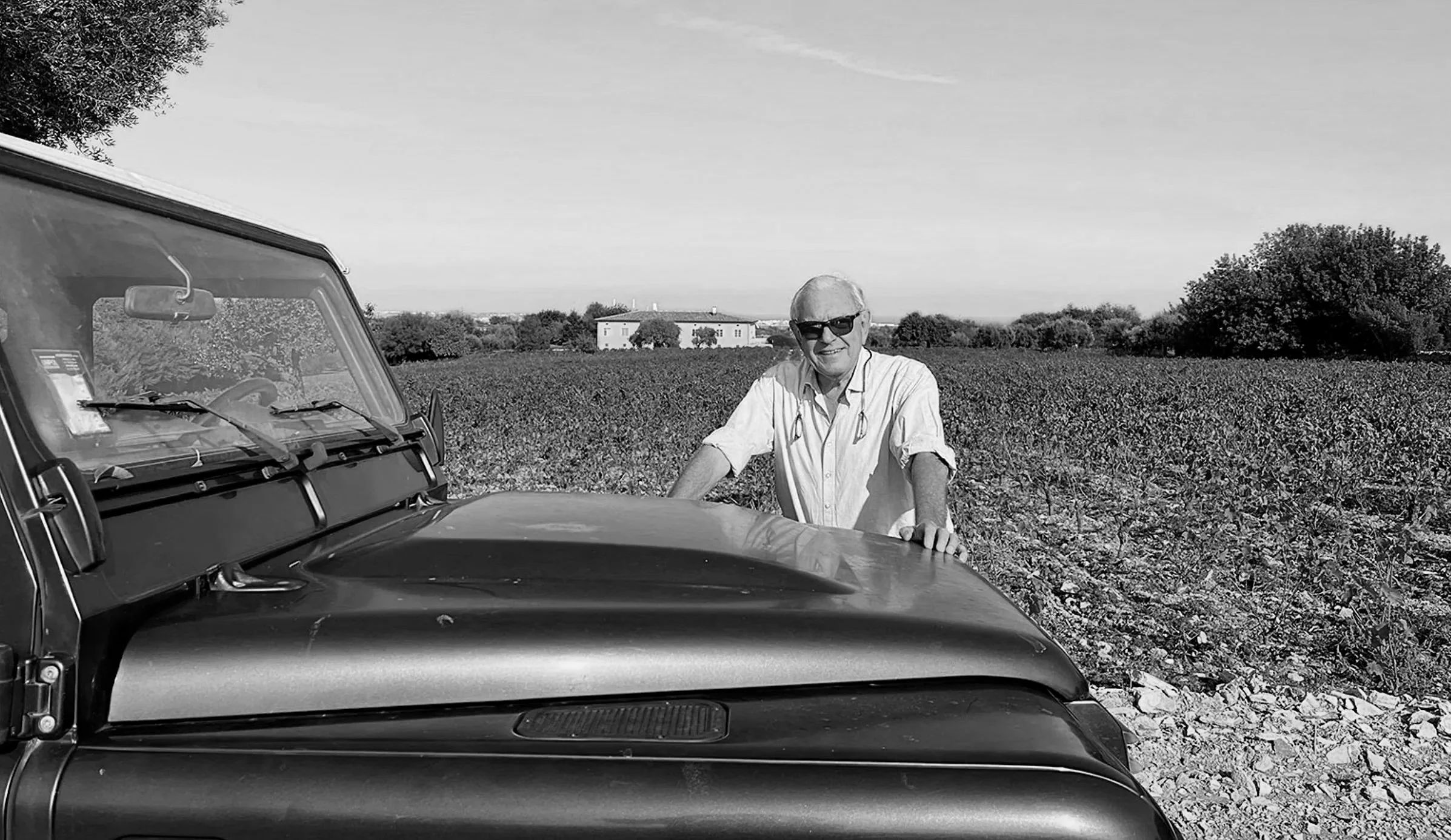 A black and white photograph of an older man wearing sunglasses, a collared shirt, and a stethoscope around his neck, standing next to a vintage car in a rural area with a cultivated field, trees, and a building in the background.
