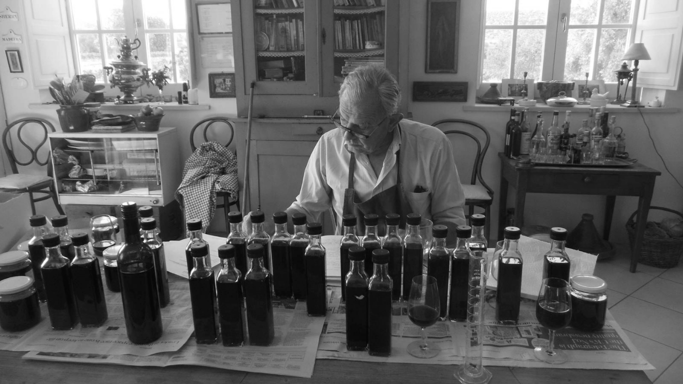 A man with white hair, glasses, and a white shirt, wearing an apron, sitting at a table with several bottles and glasses, in a room with large windows, shelves, and various containers.