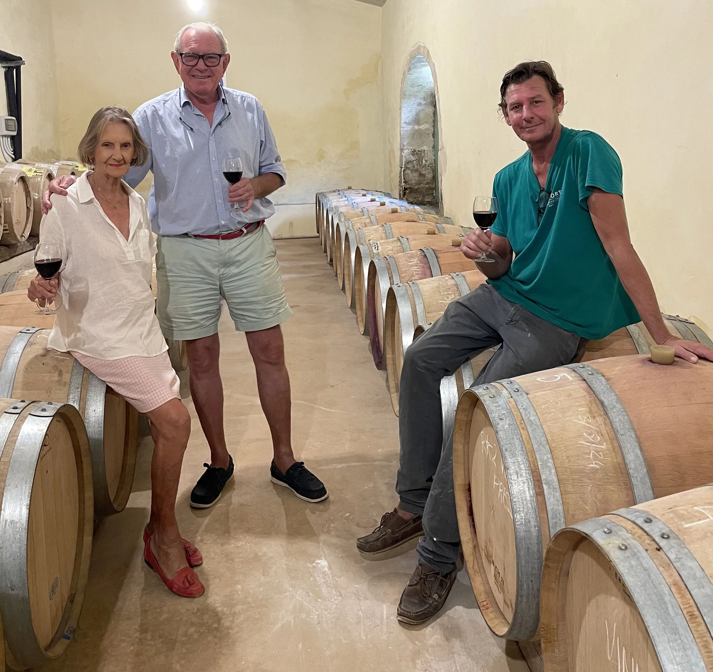 Four people inside a wine cellar, standing next to wine barrels, holding glasses of red wine, smiling at the camera.