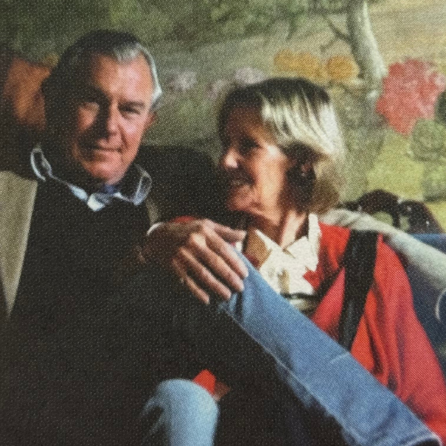 A man and a woman sitting close together, with the man smiling broadly and the woman looking towards the camera, both in casual outdoor clothing.
