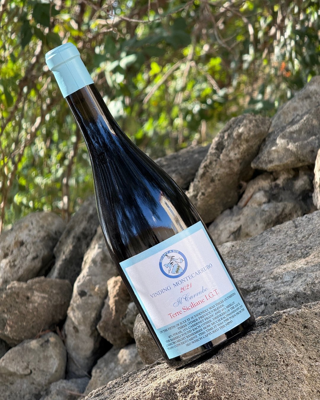 A bottle of wine resting on rocks outdoors. The label indicates it is a 2021 Terre Siciliane I.G.T. from Vinding Montecerruto, with a blue and white color scheme.