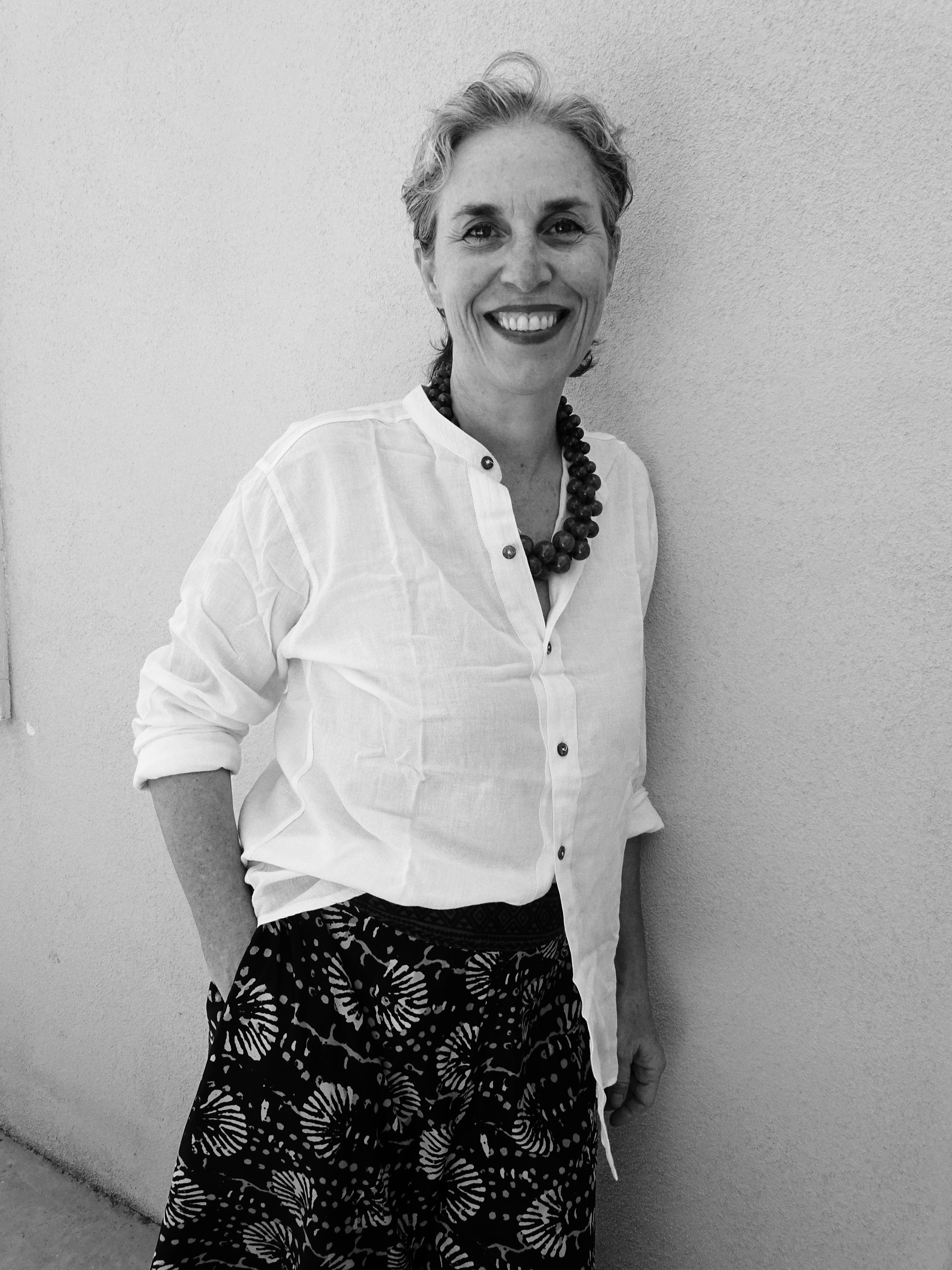 A woman with short, wavy hair smiling, wearing a light-colored button-up shirt, a large beaded necklace, and a patterned skirt, standing against a plain wall.