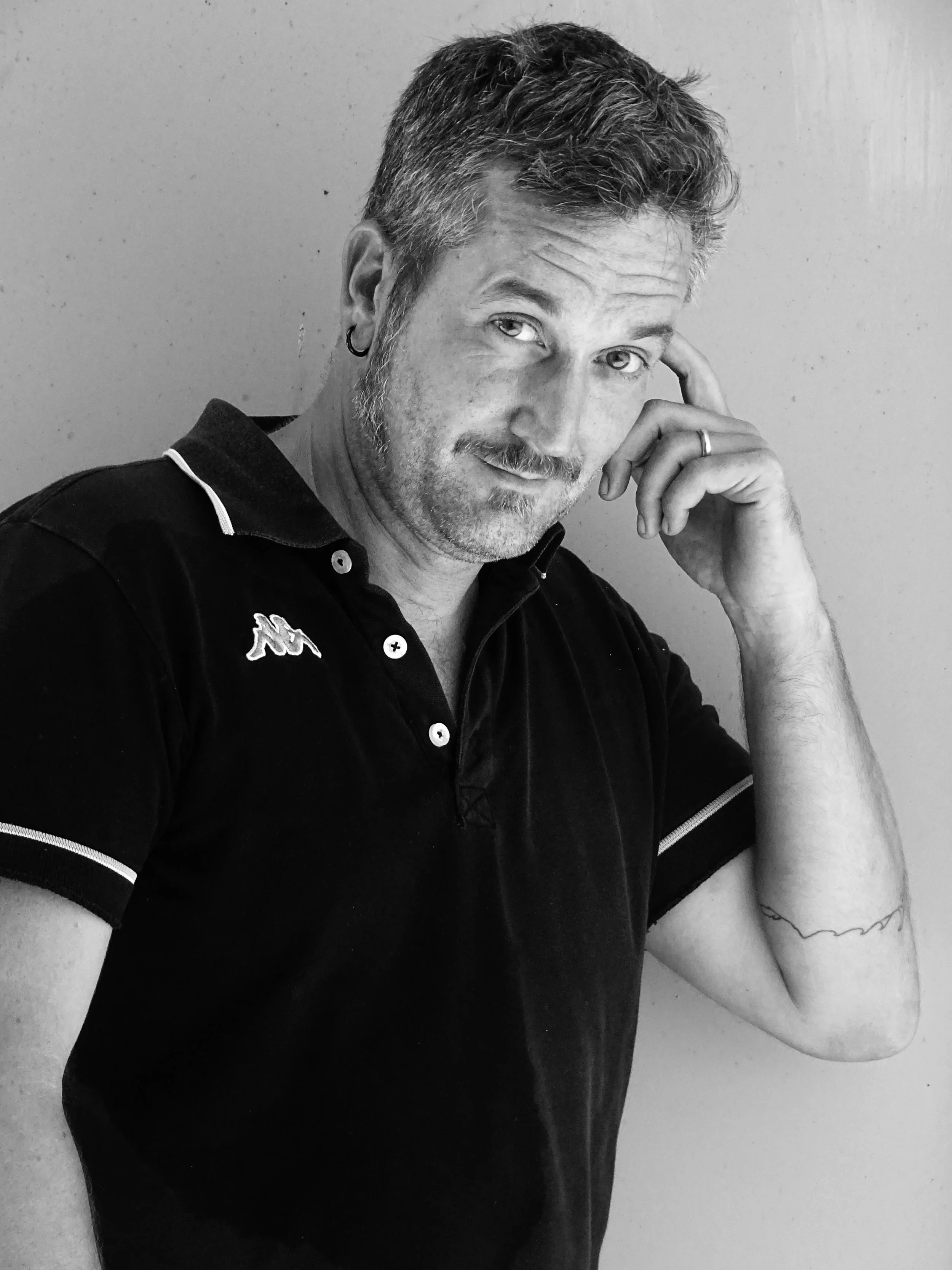 A black and white photo of a man with short, curly hair, a beard, and an earring in his left ear. He is wearing a dark collared shirt with a logo on the chest and a ring on his left hand. He is resting his chin on his left hand and looking at the camera with a slight smile, standing against a plain wall.