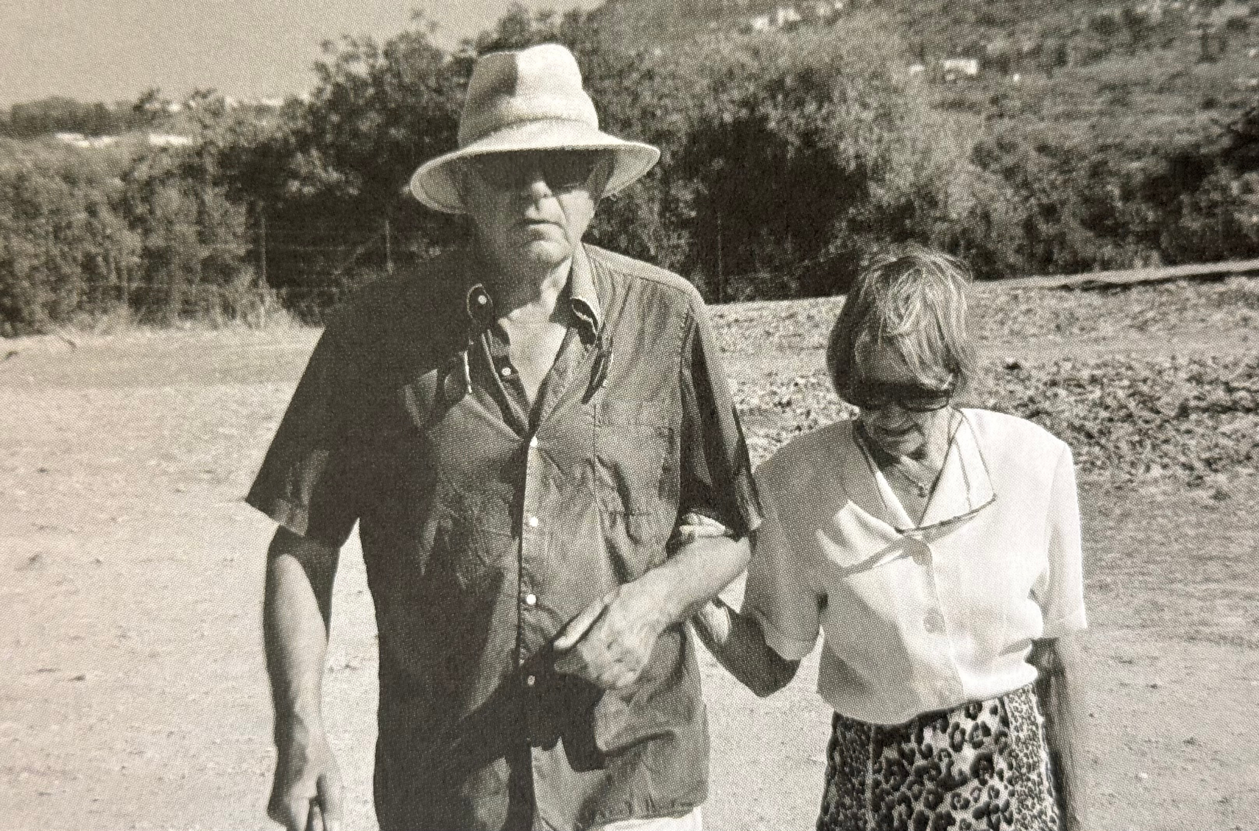 Black-and-white photo of a man in a straw hat and a woman with a short haircut holding hands outdoors, with trees and a pathway in the background.