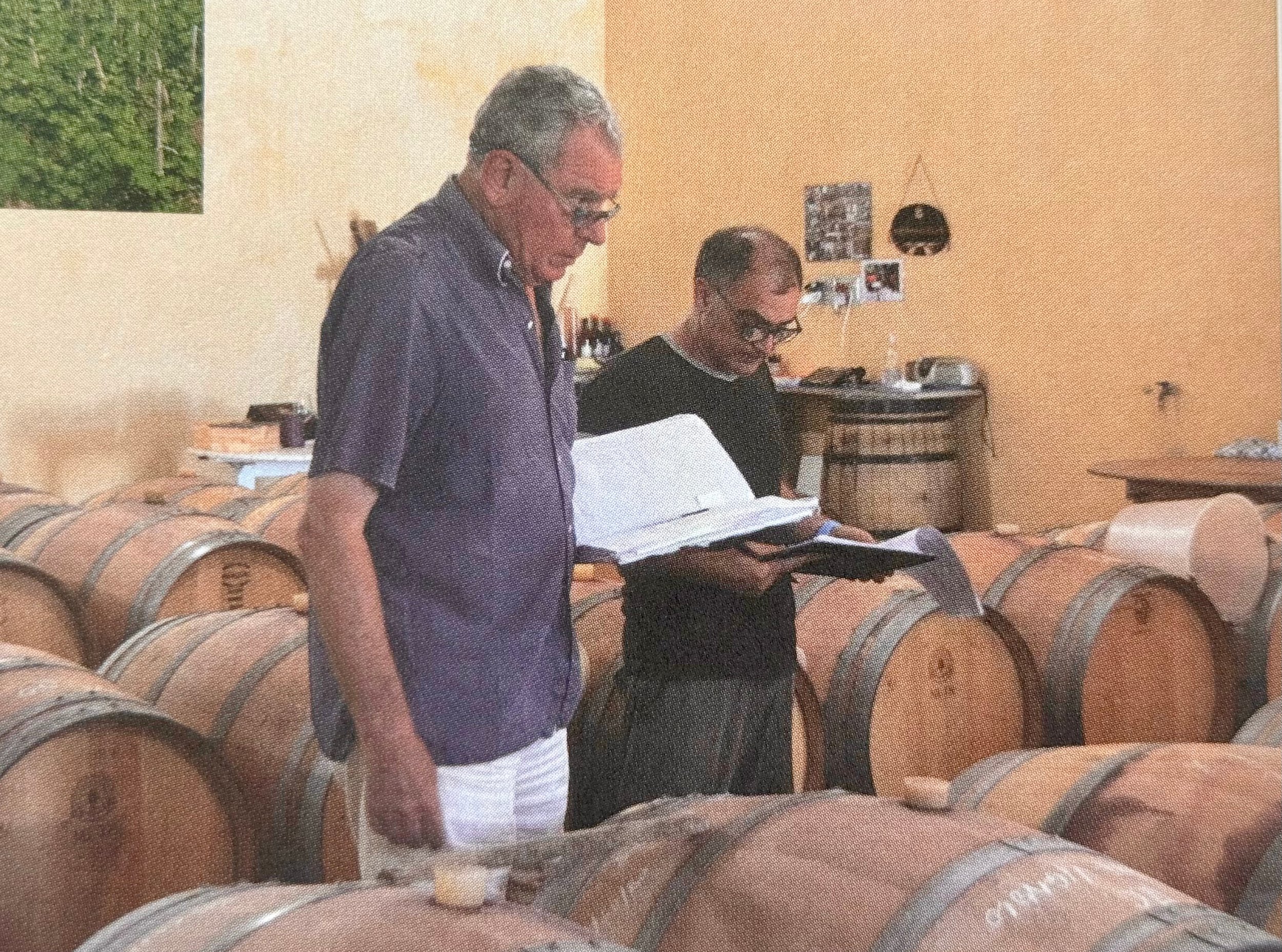 Two men are standing in a room filled with wine barrels, examining a chart or book.