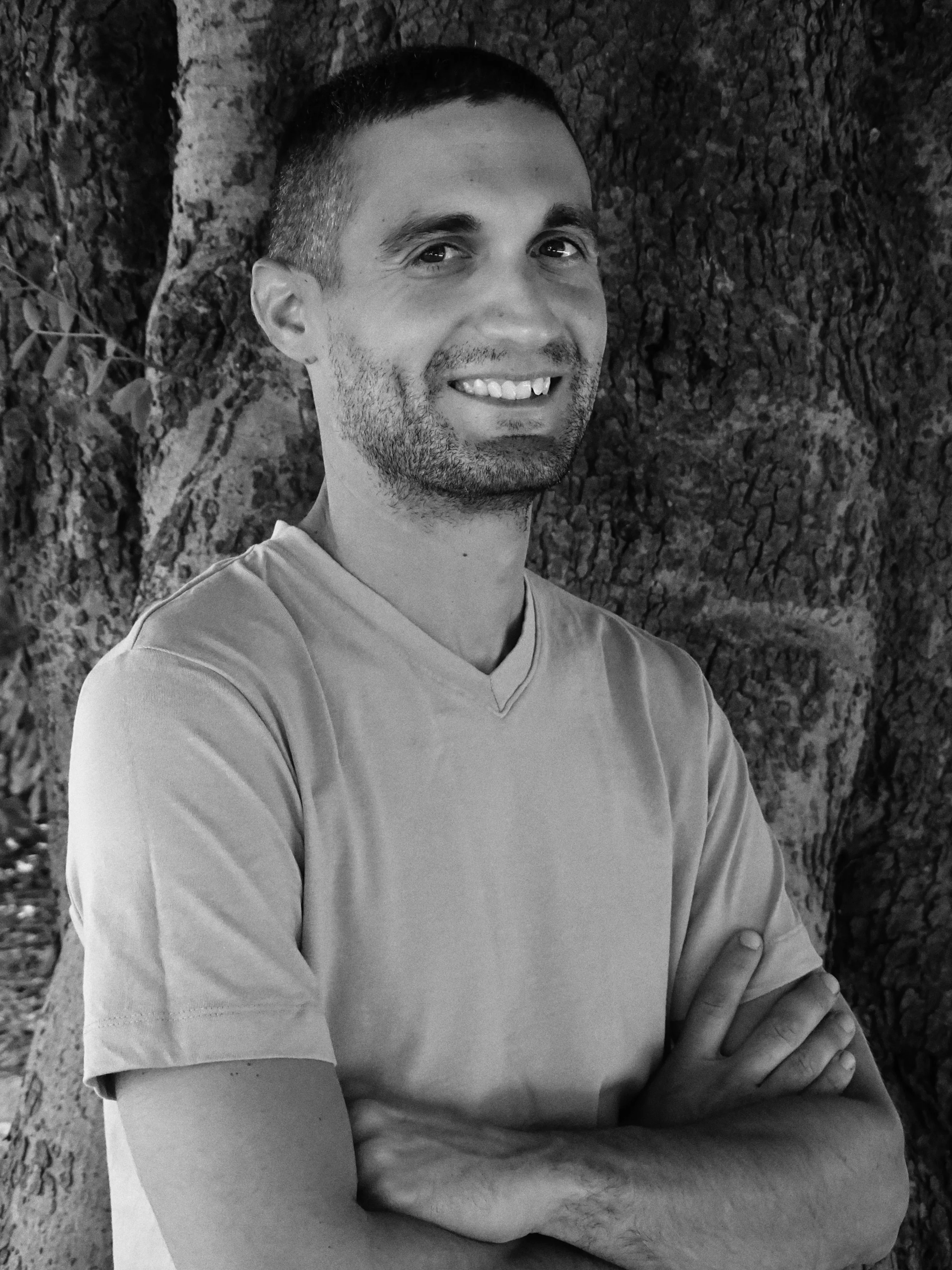 Black and white portrait of a smiling man with short haircut and a beard, standing outdoors with a large tree trunk in the background, arms crossed in front of him.