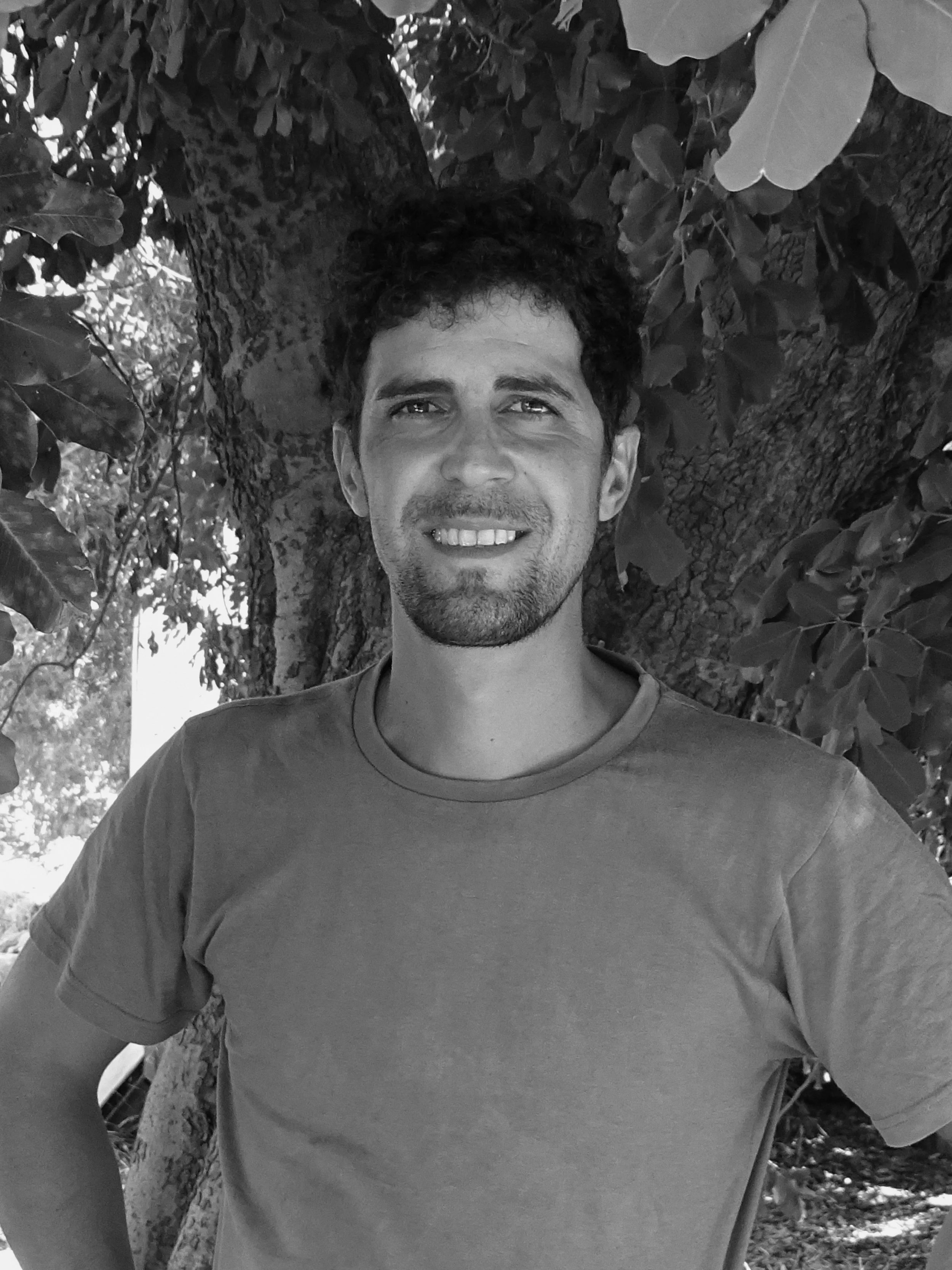 Close-up of a smiling man standing outdoors next to a tree with leaves, partial view of his face and upper body visible in black and white.