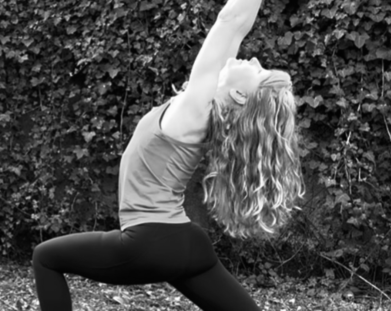 A woman practicing yoga outdoors in front of a leafy fence, performing a pose with one arm raised overhead and her face tilted upward.