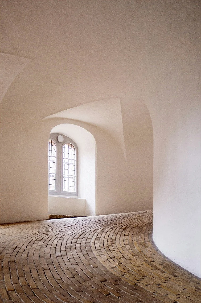 Empty interior space with curved white walls, a large arched window with grid panes, and a arched brick floor.