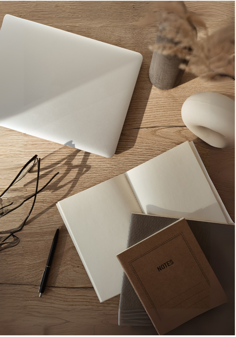 A top-down view of a wooden desk with a closed silver laptop, an open notebook, a brown notepad labeled "NOTES", a pair of glasses, a black pen, a ceramic vase with dried flowers, a white sculptural object, and a blank card.
