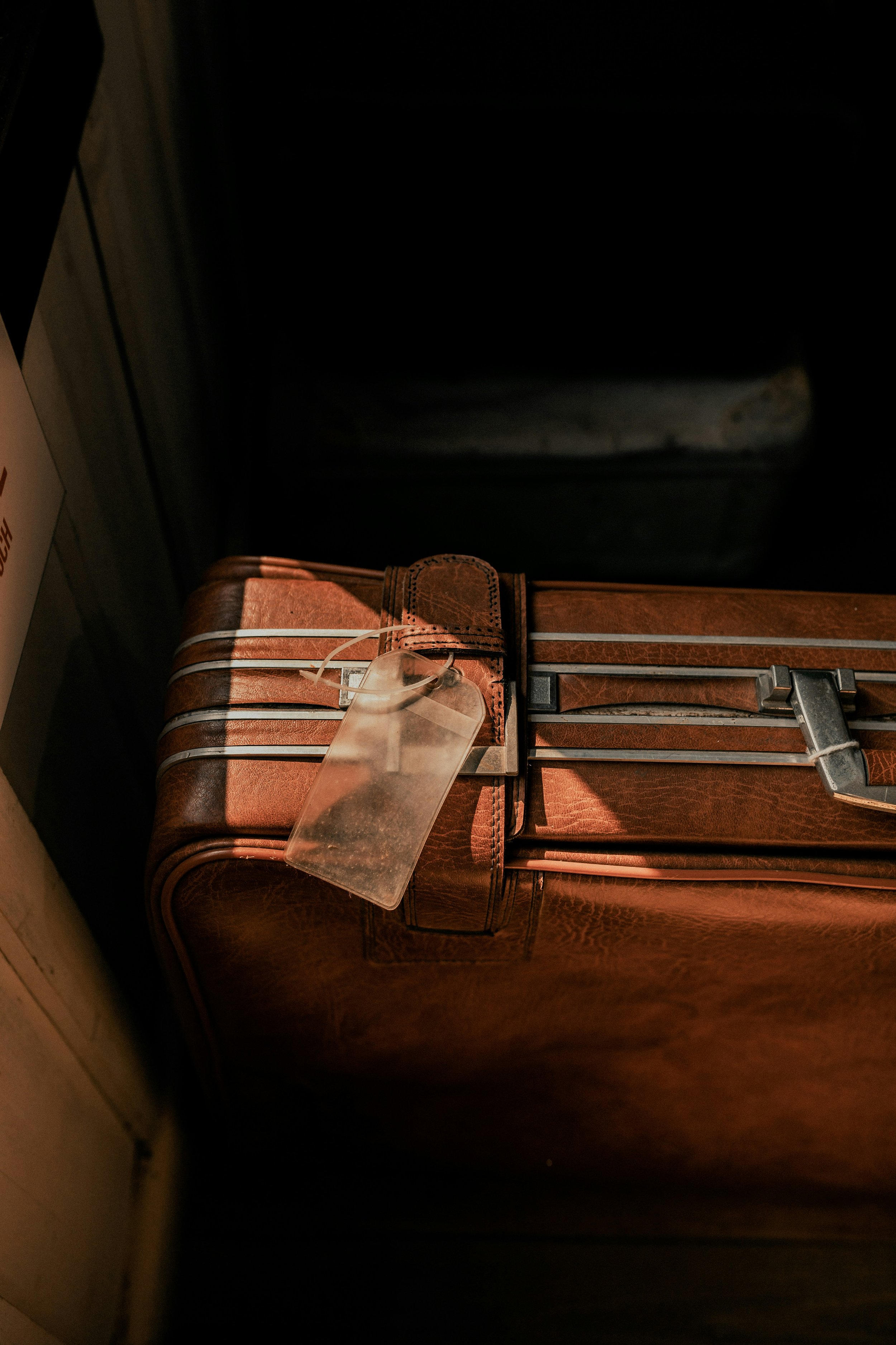 A close-up picture of a brown leather suitcase with metal buckles, secured with a strap, with a clear plastic luggage tag attached, placed on a flat surface.