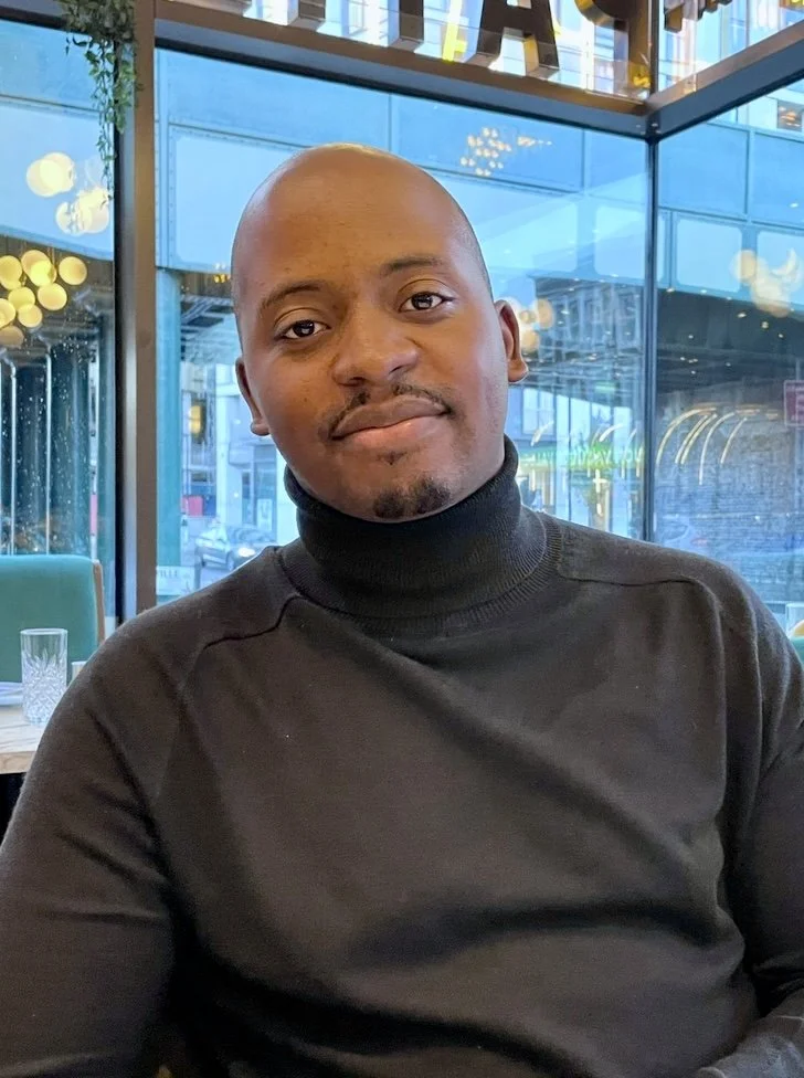 A man with a shaved head and a goatee, wearing a black turtleneck sweater, sitting indoors near large glass windows with city street view in the background.