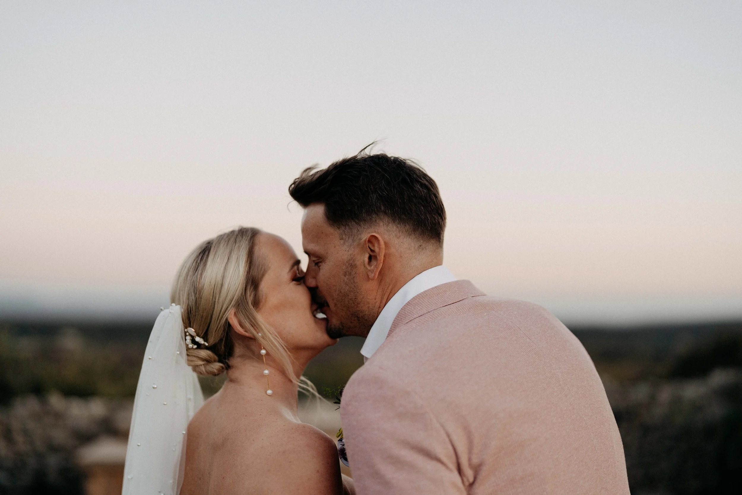 A bride and groom are kissing outdoors during sunset or dusk, with the sky in the background.