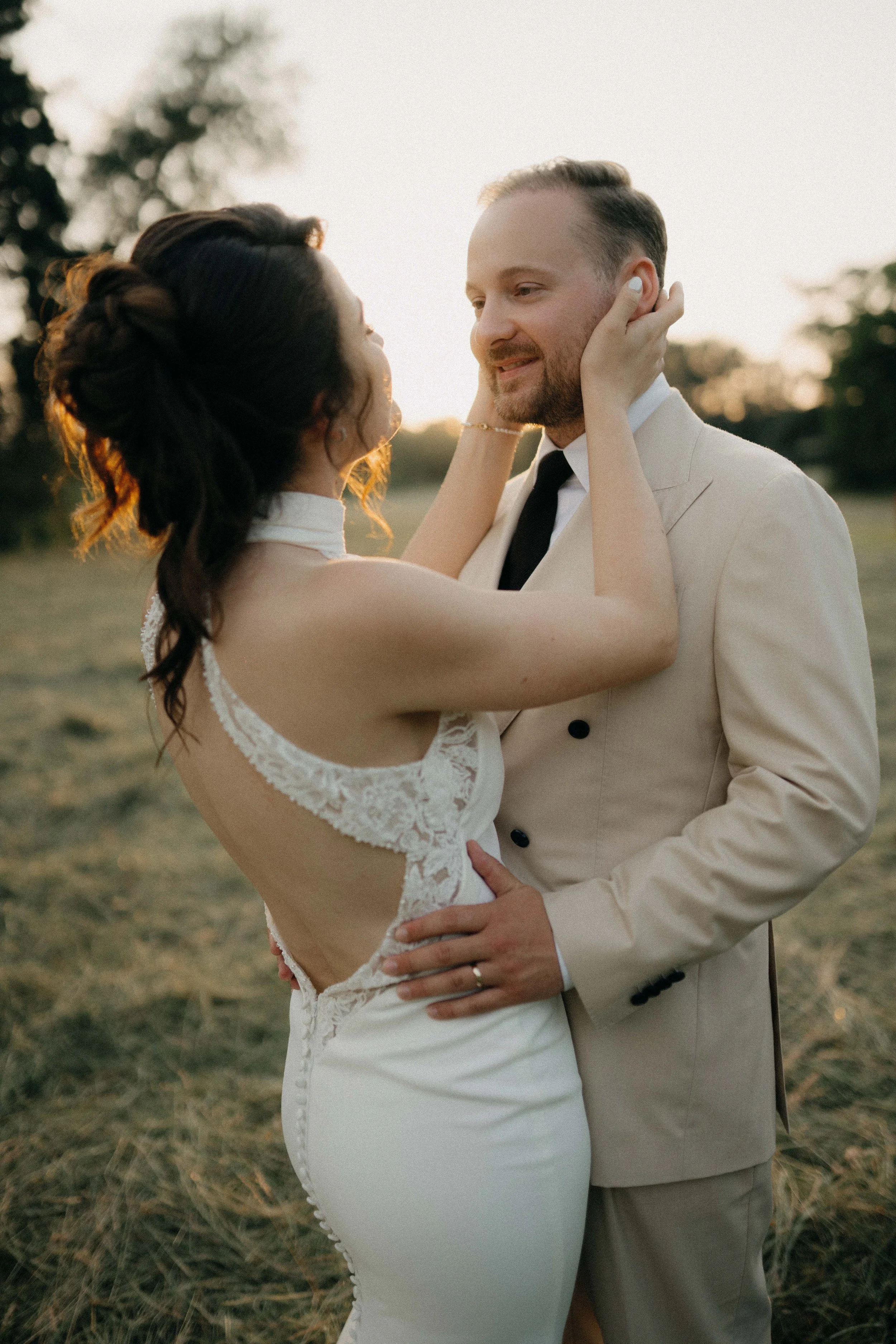 A bride and groom holding each other closely outdoors during sunset, with the bride's hands on the groom's face, both gazing into each other's eyes.