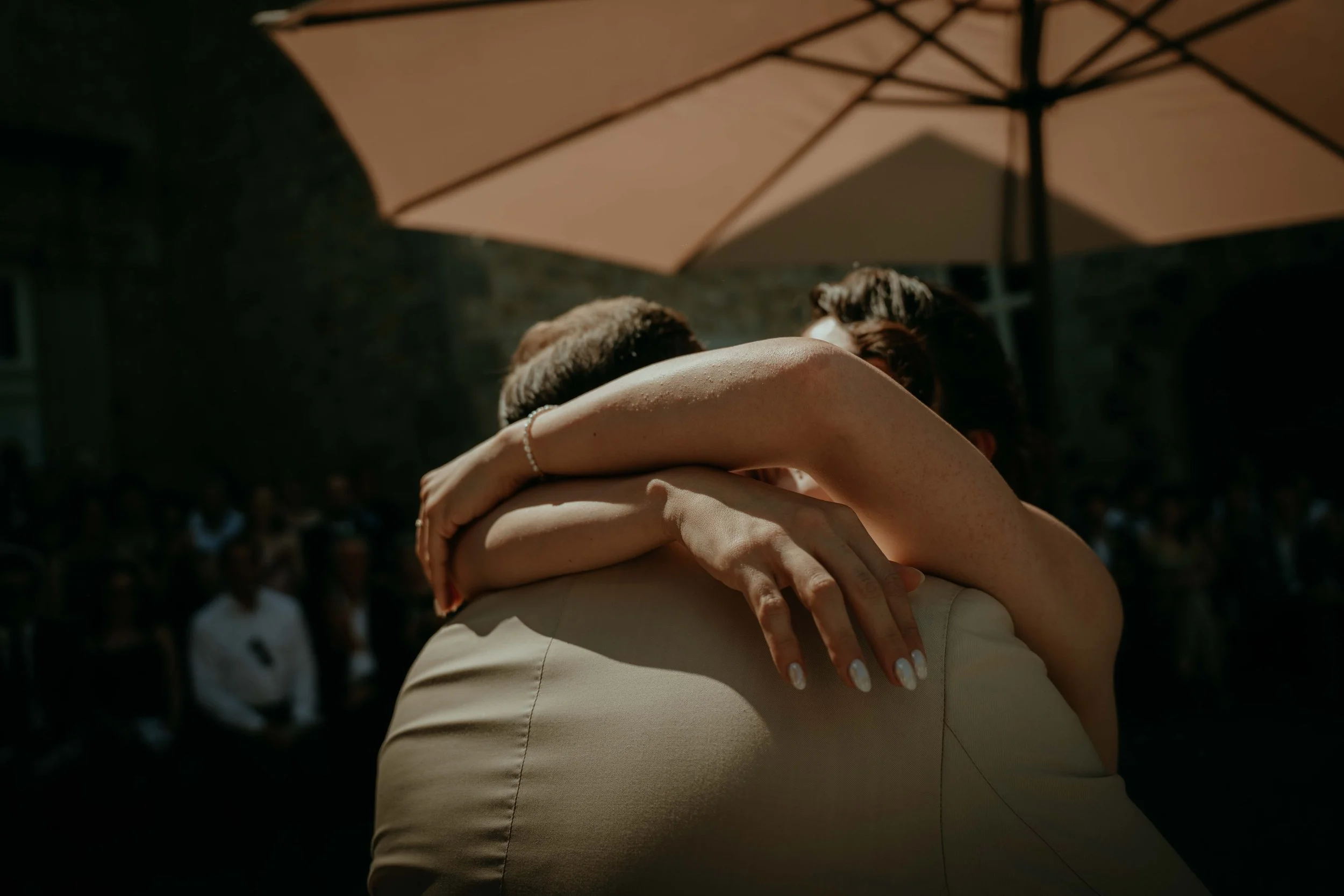 Two people embracing each other at an outdoor event with a large umbrella overhead. The scene is warm and intimate, with blurred audience in the background.