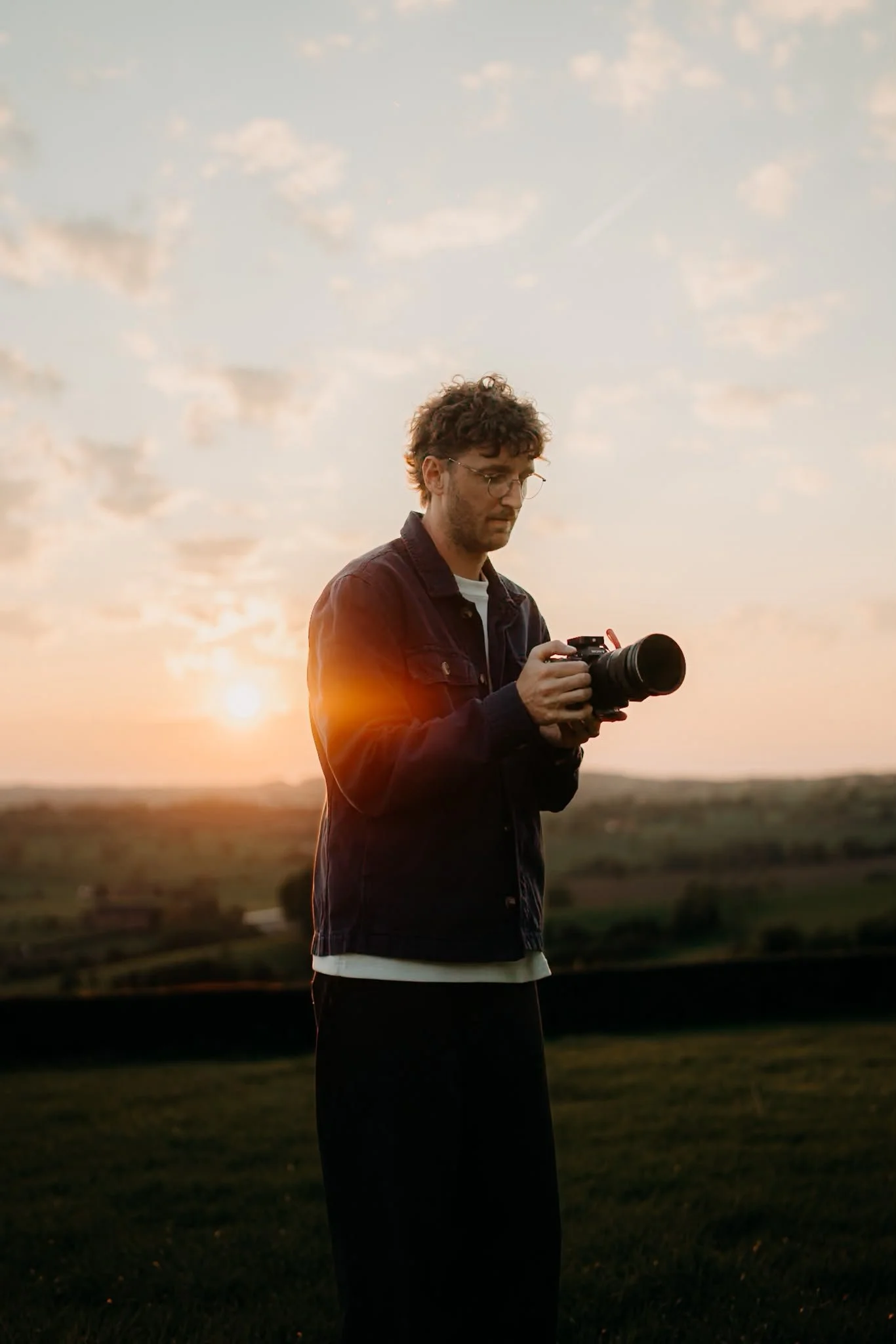 A man with curly hair and glasses stands outdoors at sunset, looking at his camera, with a landscape and sky with clouds in the background.