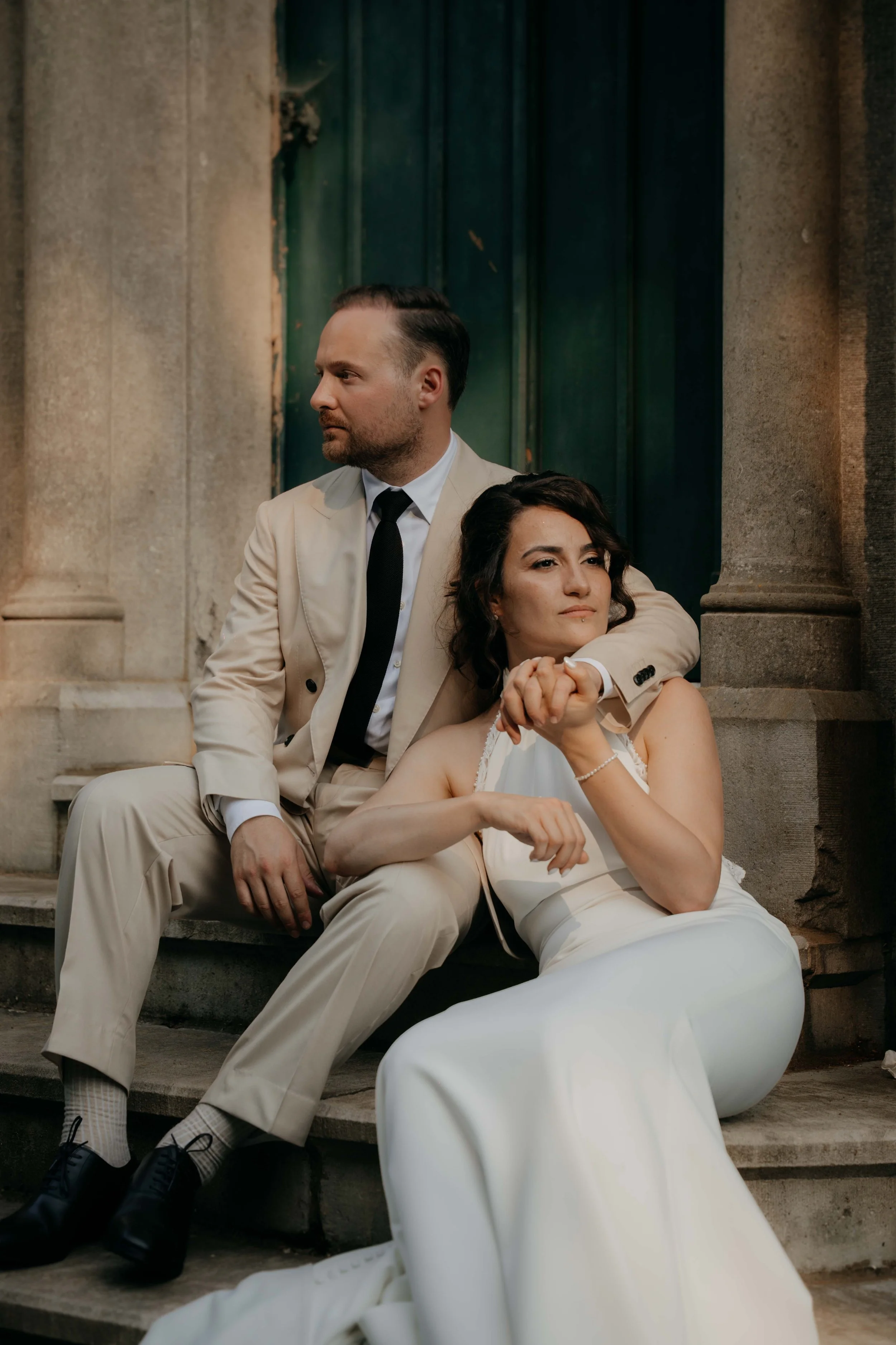 A man in a beige suit and tie sitting on steps with a woman in a white dress leaning on his shoulder, both in front of a stone building with columns and dark wooden doors.
