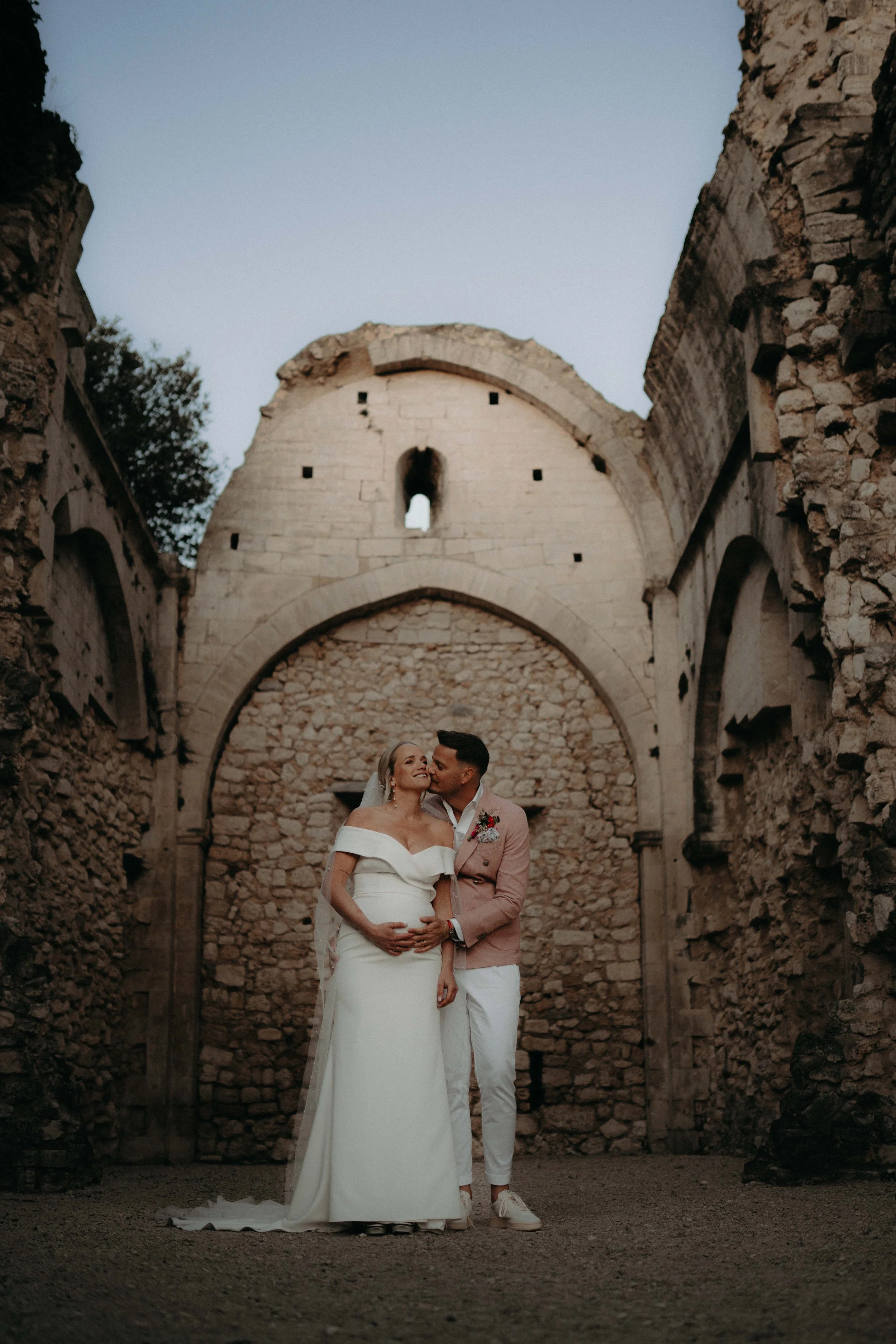 A bride and groom standing together in front of an old stone structure with arches, during their wedding photoshoot at dusk.