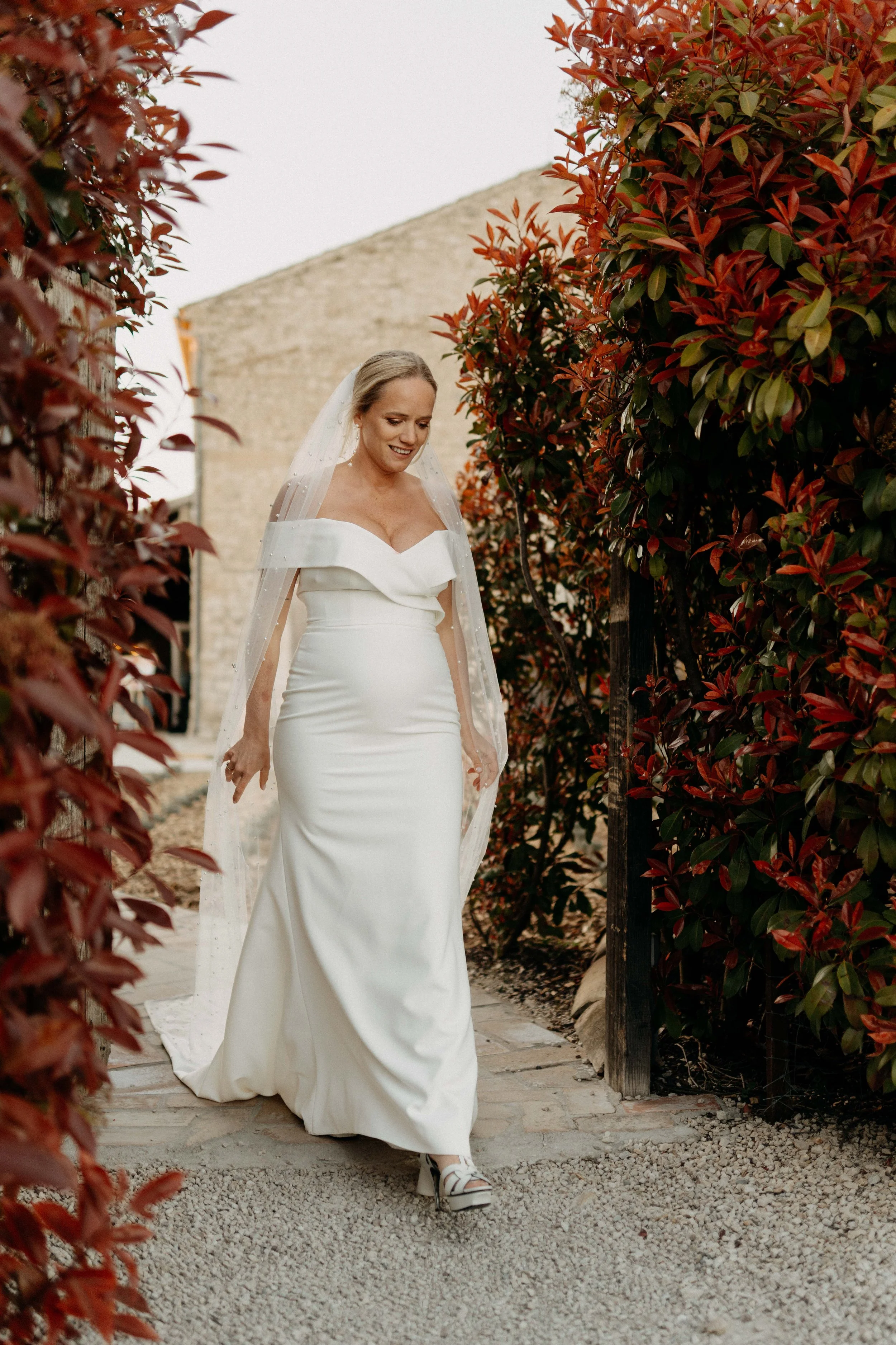 A bride in a white wedding gown walking outdoors between red and green foliage with a brick building in the background.