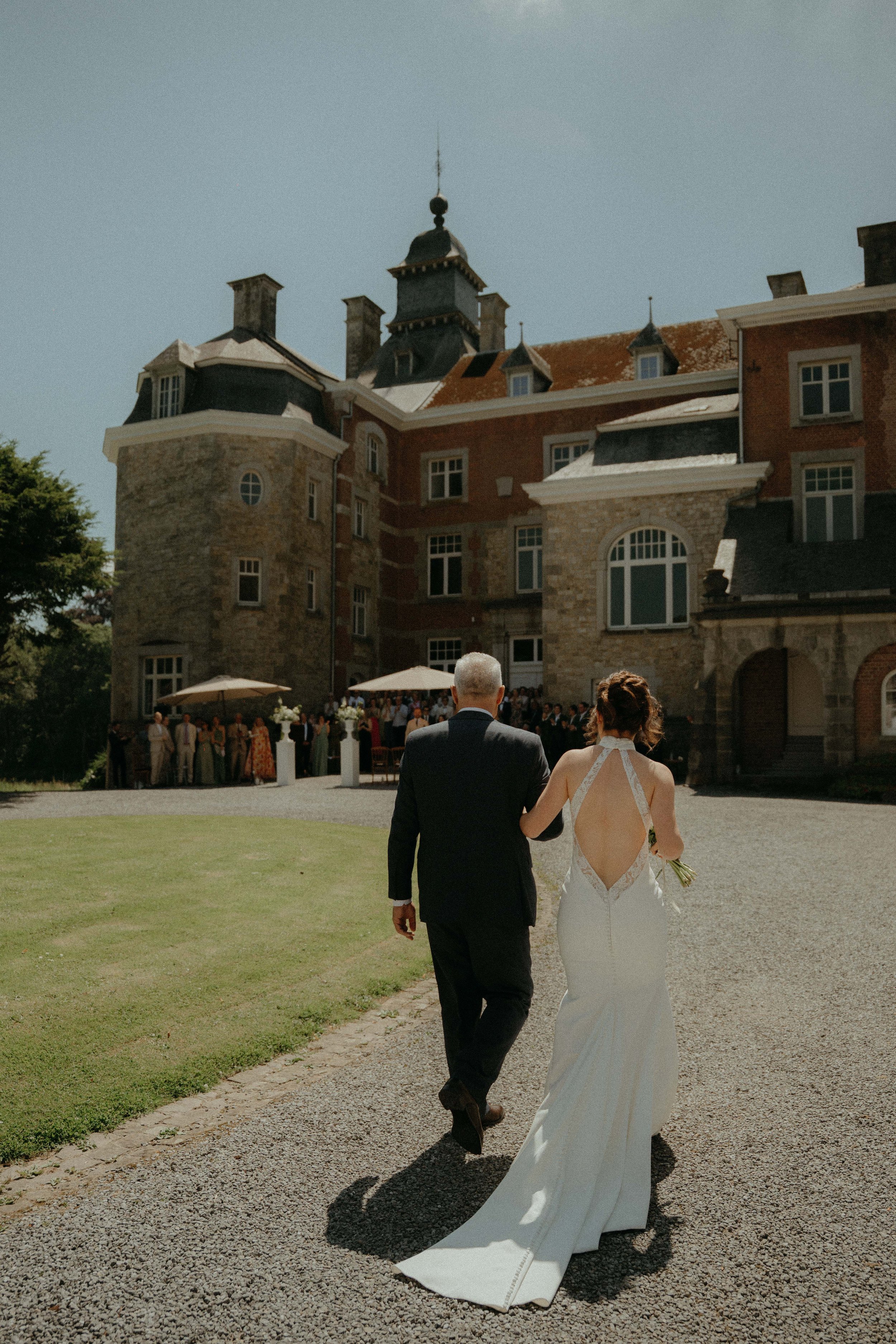 A newlywed couple walking towards a historic mansion with a courtyard filled with guests under blue skies.