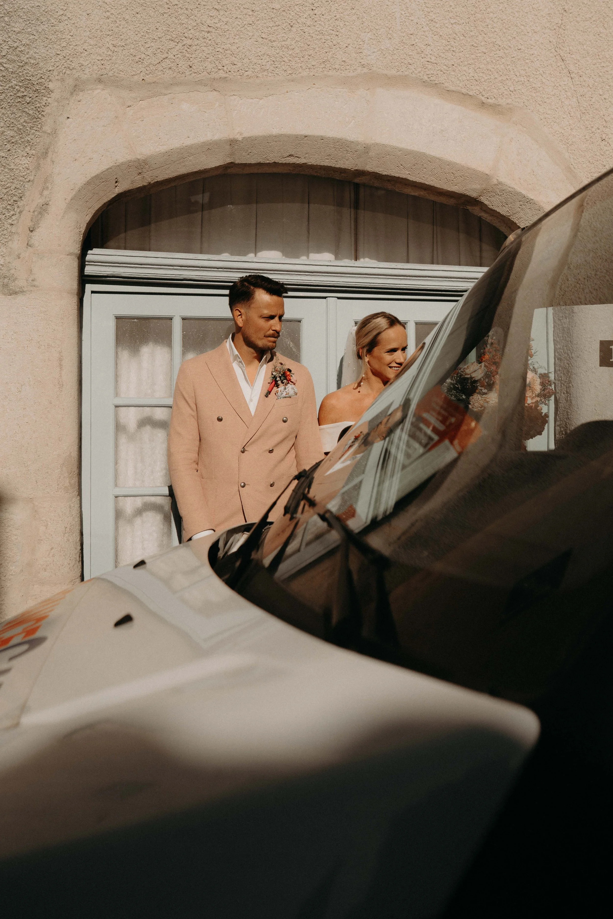 A bride and groom standing outside a building in front of a door with a large arched window above it, while a police car is parked in front of them, reflecting their image on its hood.