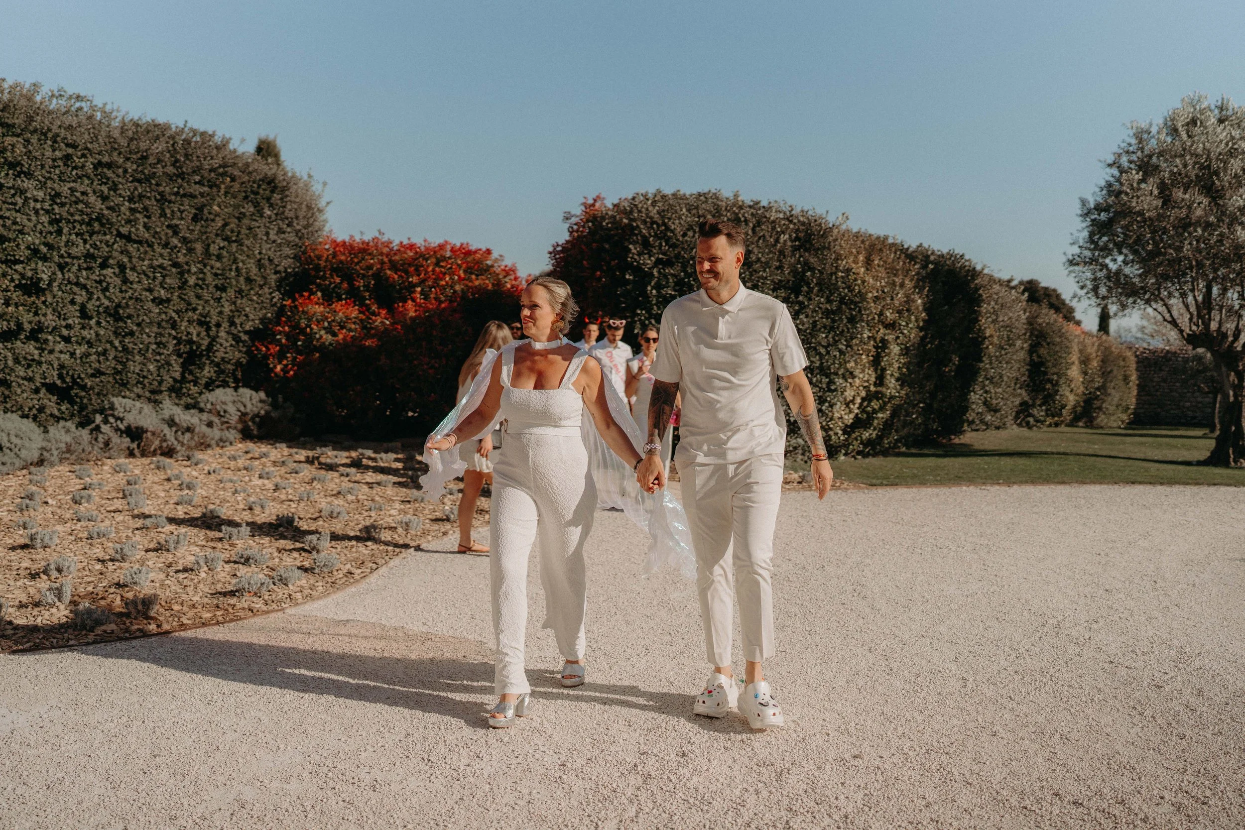 A group of people walking outdoors in a park, two individuals in foreground holding hands, a man and a woman in white clothing, smiling and enjoying a sunny day, with large bushes and trees in the background.