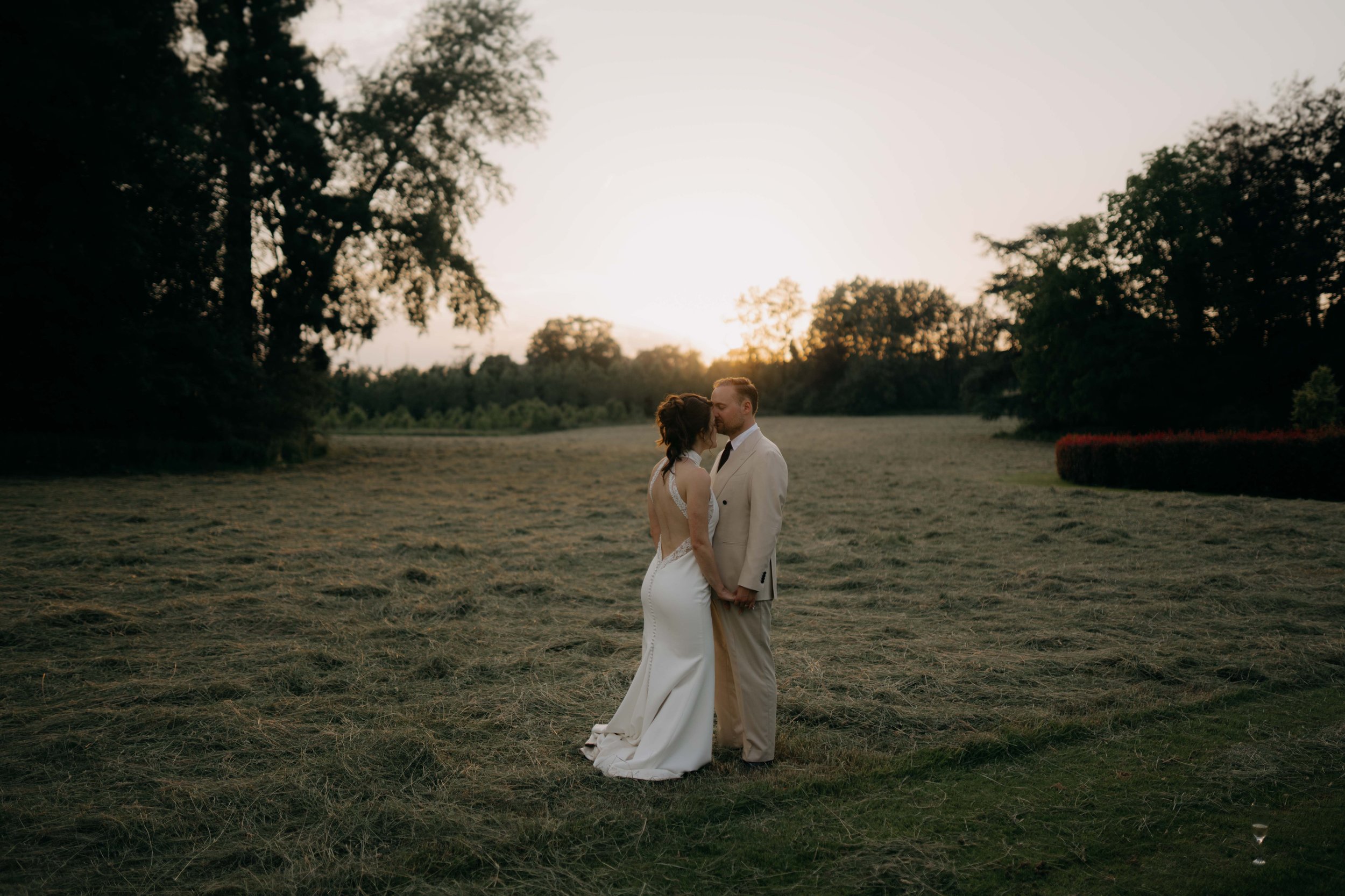 A couple dressed in wedding attire, standing on a grassy field with trees in the background and the sun setting, sharing a kiss.