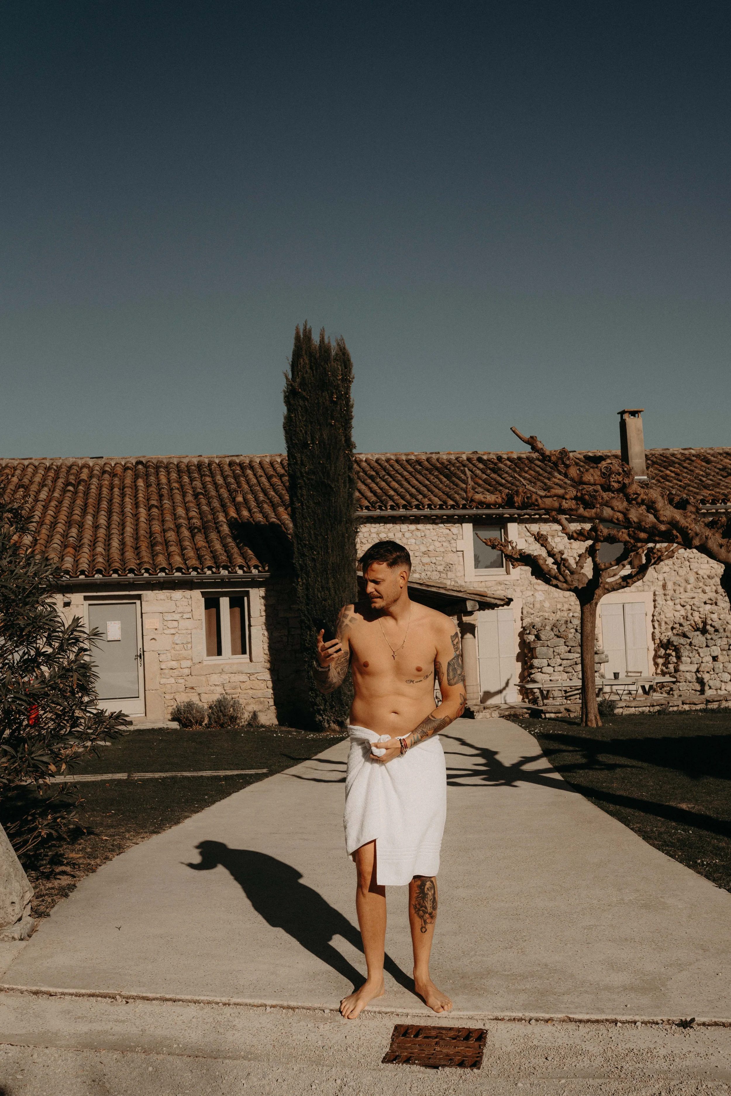 A shirtless man with tattoos, wearing a white towel around his waist, standing barefoot outdoors on a concrete pathway in front of a house with a tiled roof, trees, and bushes, holding a towel and looking at his phone.