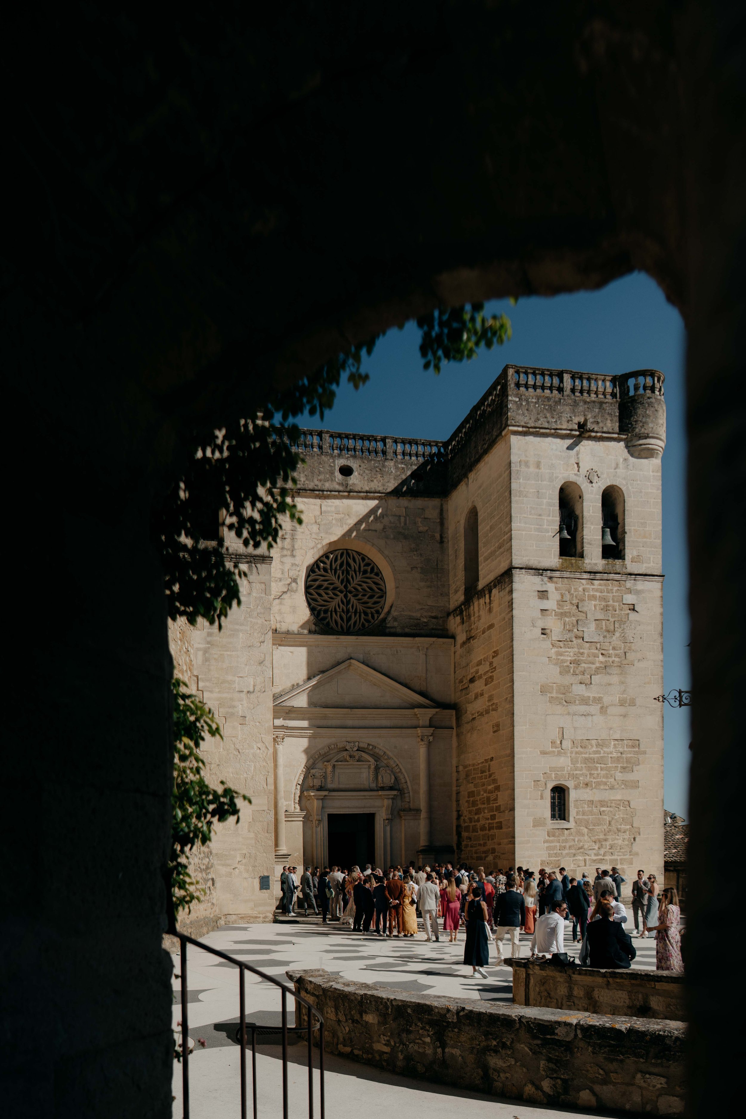 A large stone church with a crowd of people gathered at the entrance, seen through an archway with ivy.