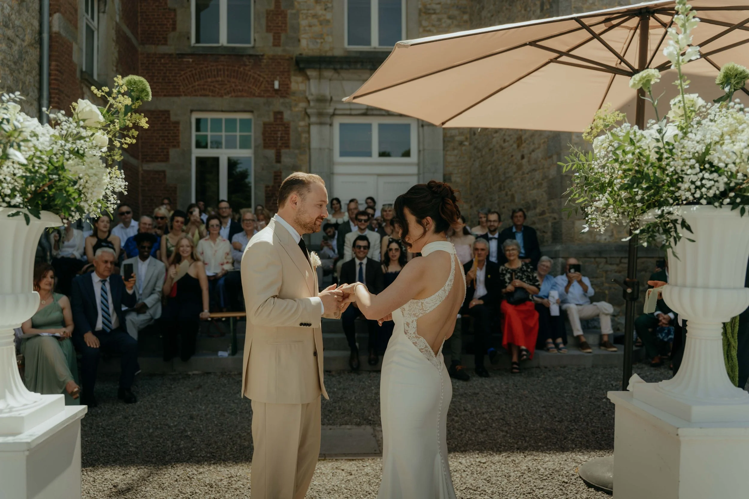 A bride and groom exchanging vows at an outdoor wedding ceremony, with guests seated and watching. The ceremony is decorated with large white floral arrangements and umbrellas for shade, set against a building with stone and brick architecture.