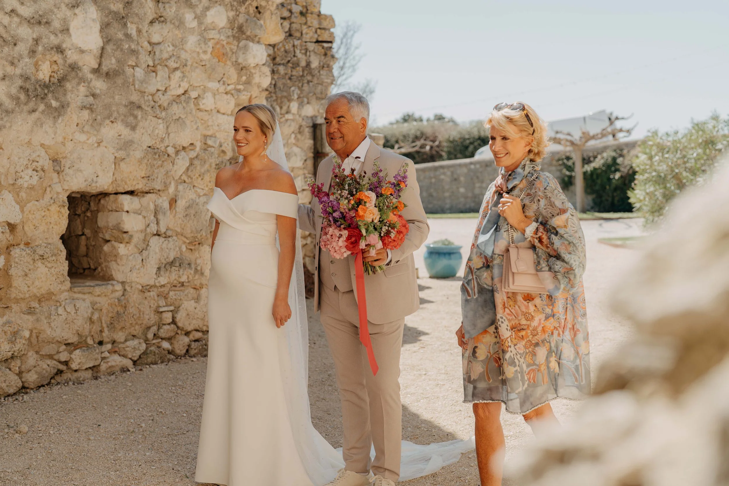 A bride in a white wedding dress, an older man holding a bouquet of flowers, and a woman in floral attire stand outdoors by a stone wall, smiling during a wedding or special event.