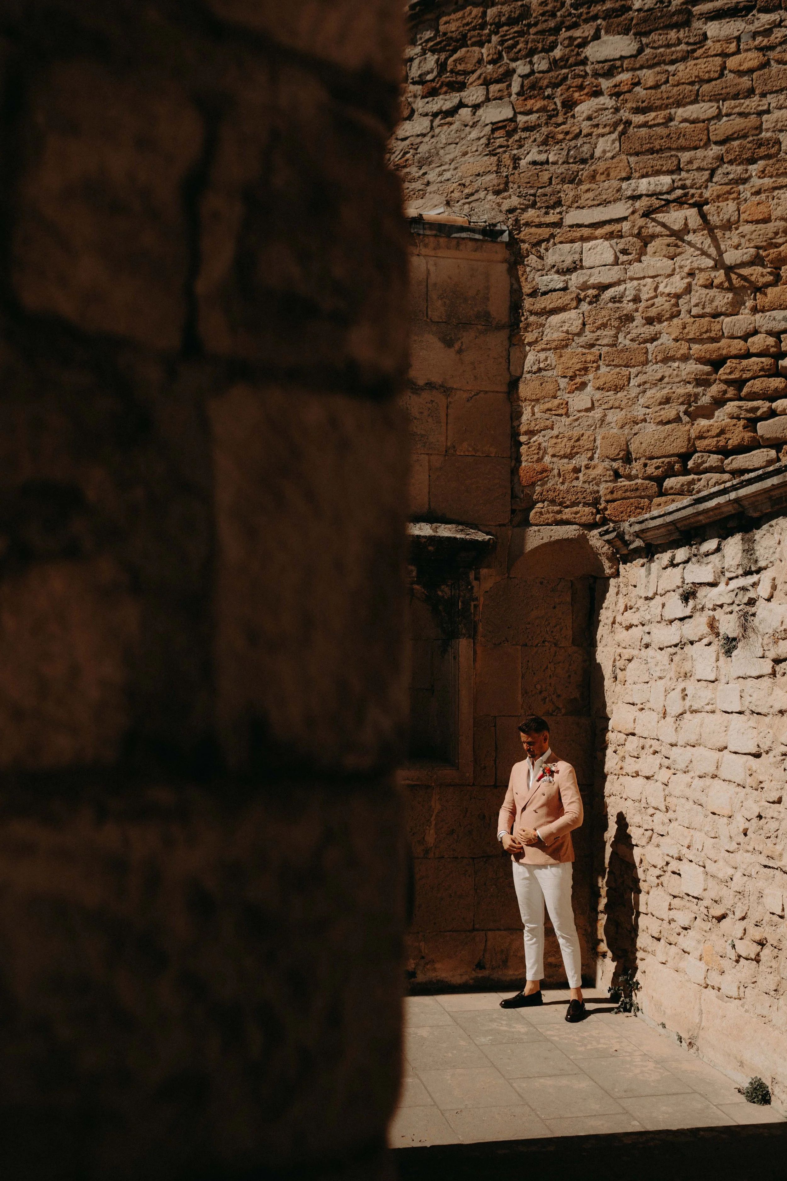 A man in a beige blazer and white pants stands against an ancient stone wall, illuminated by sunlight, with a focused expression.