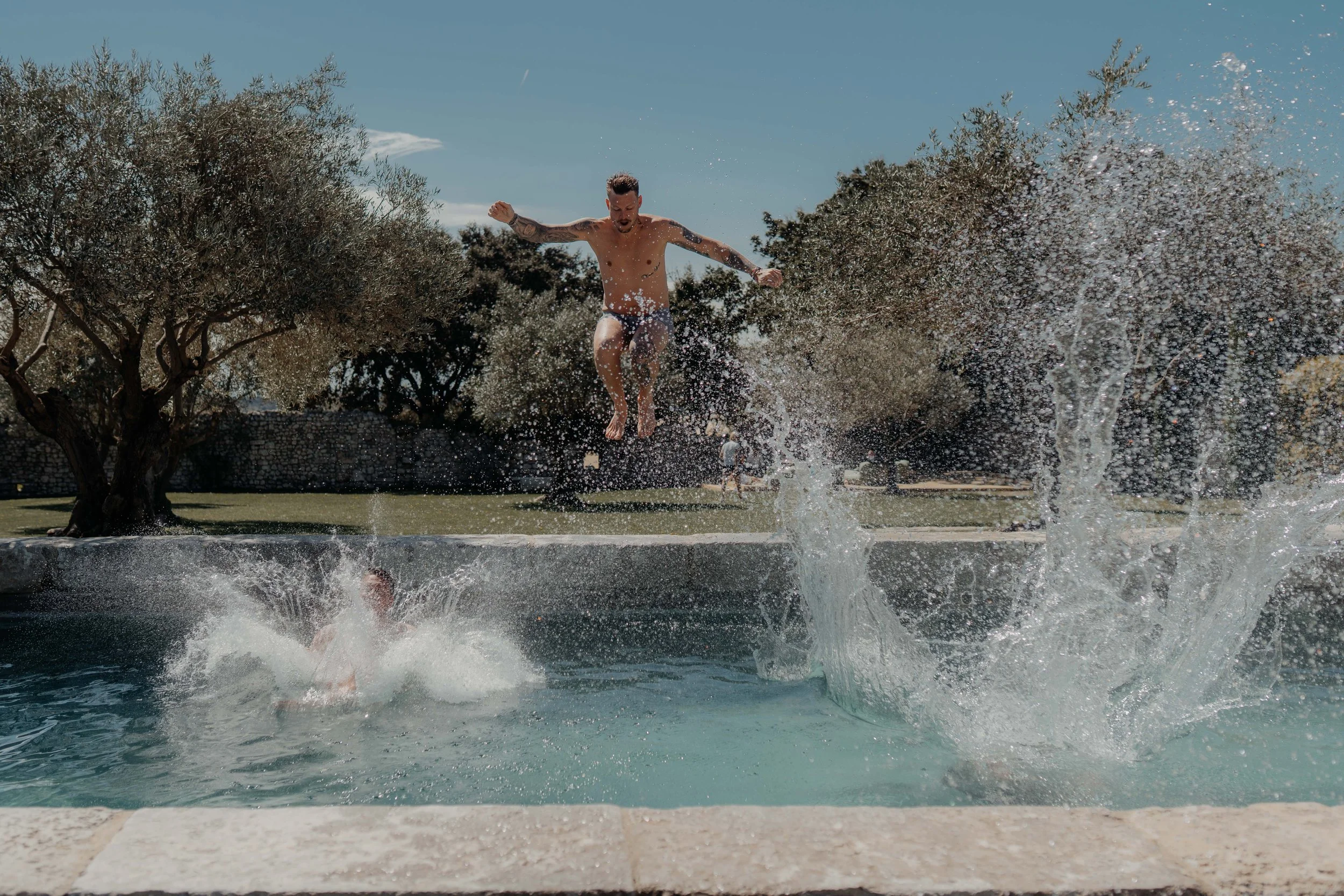 A man jumping into a pool from a concrete edge during the daytime with trees and a clear blue sky in the background, splashing water around.