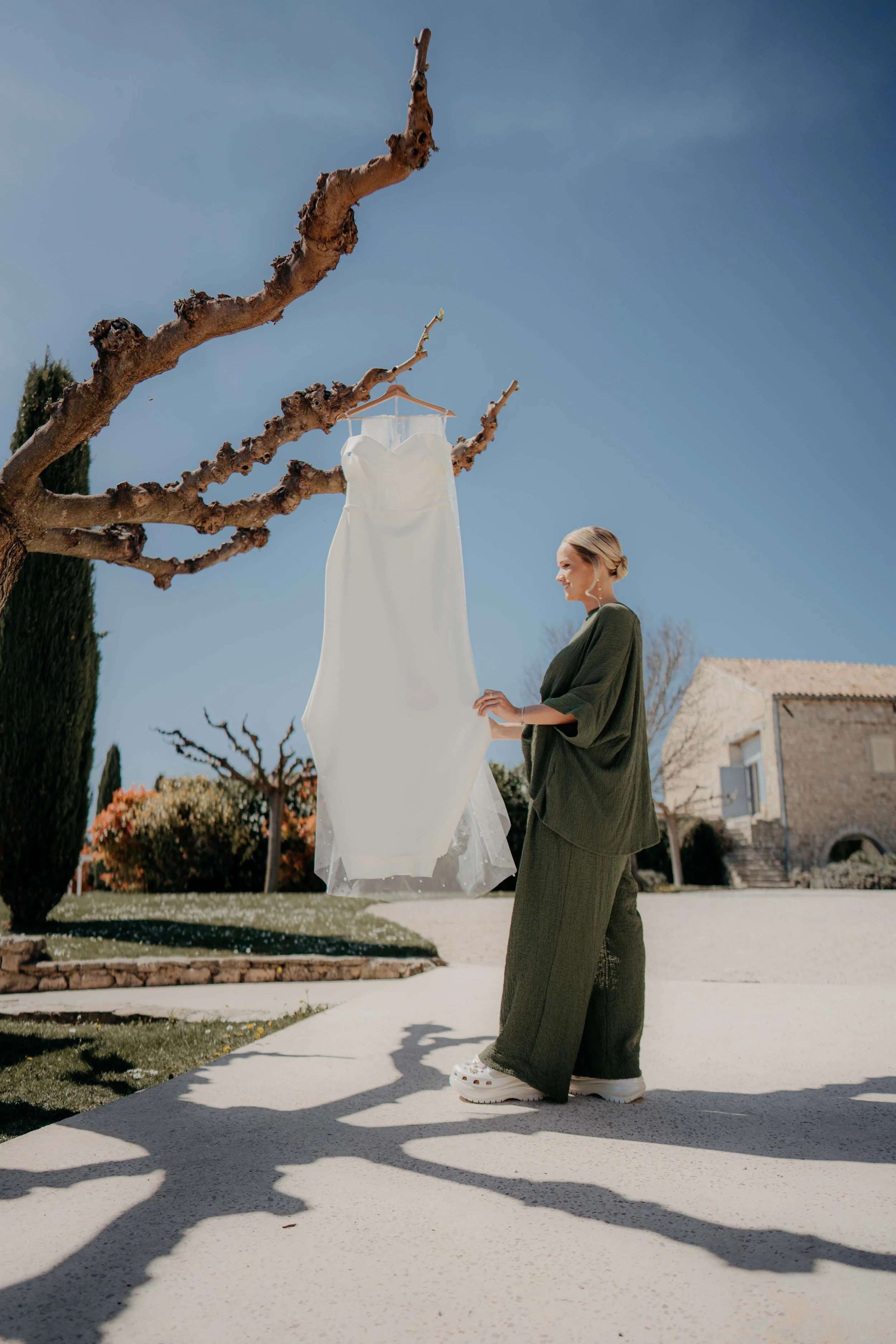 A woman in dark green baggy pants, a matching top, and white chunky sneakers stands outdoors, smiling at a white dress hanging on a branch of a leafless tree against a clear blue sky.
