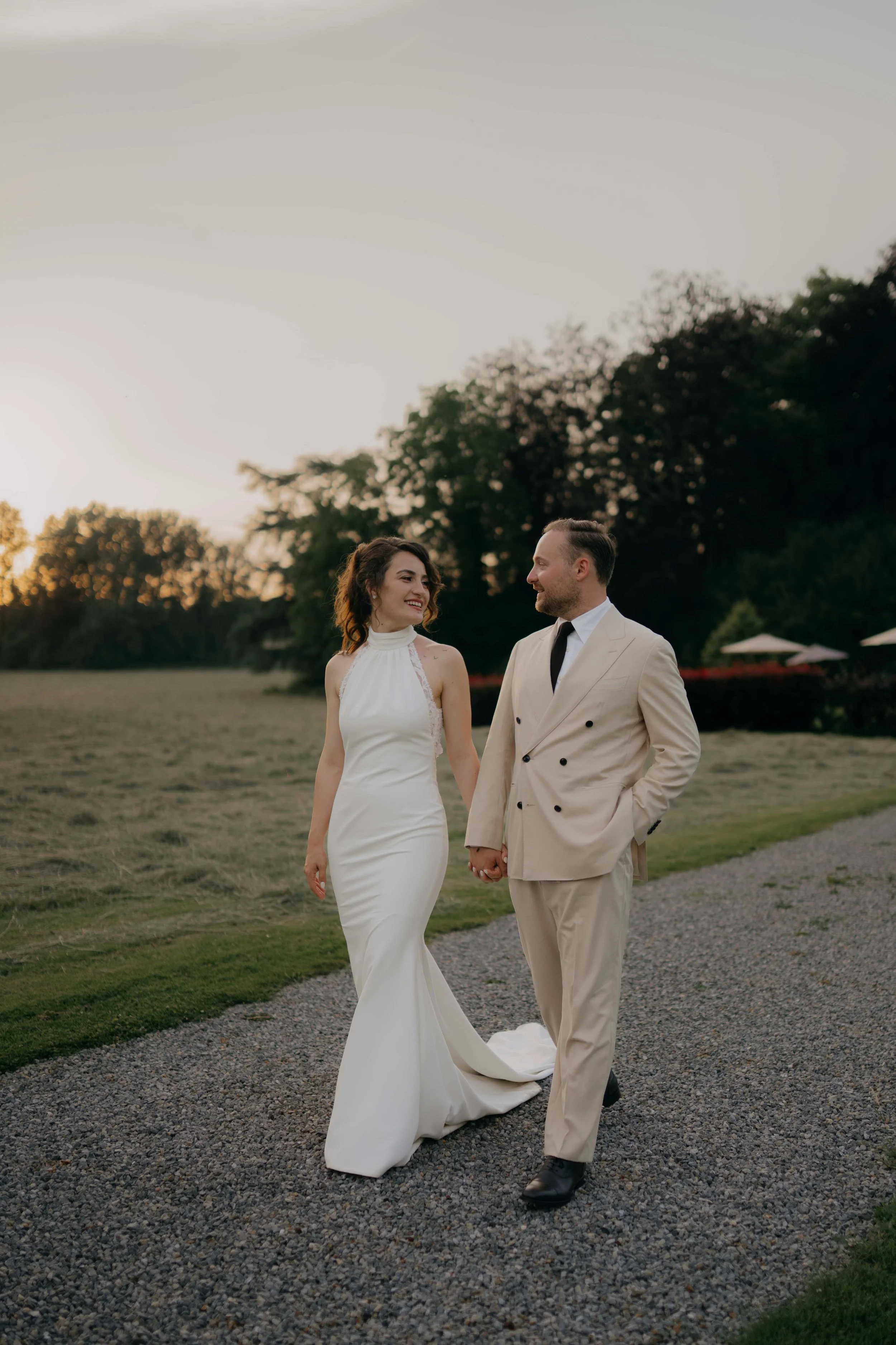 A bride and groom holding hands and walking outdoors on a gravel path at sunset, with trees and a pavilion in the background.