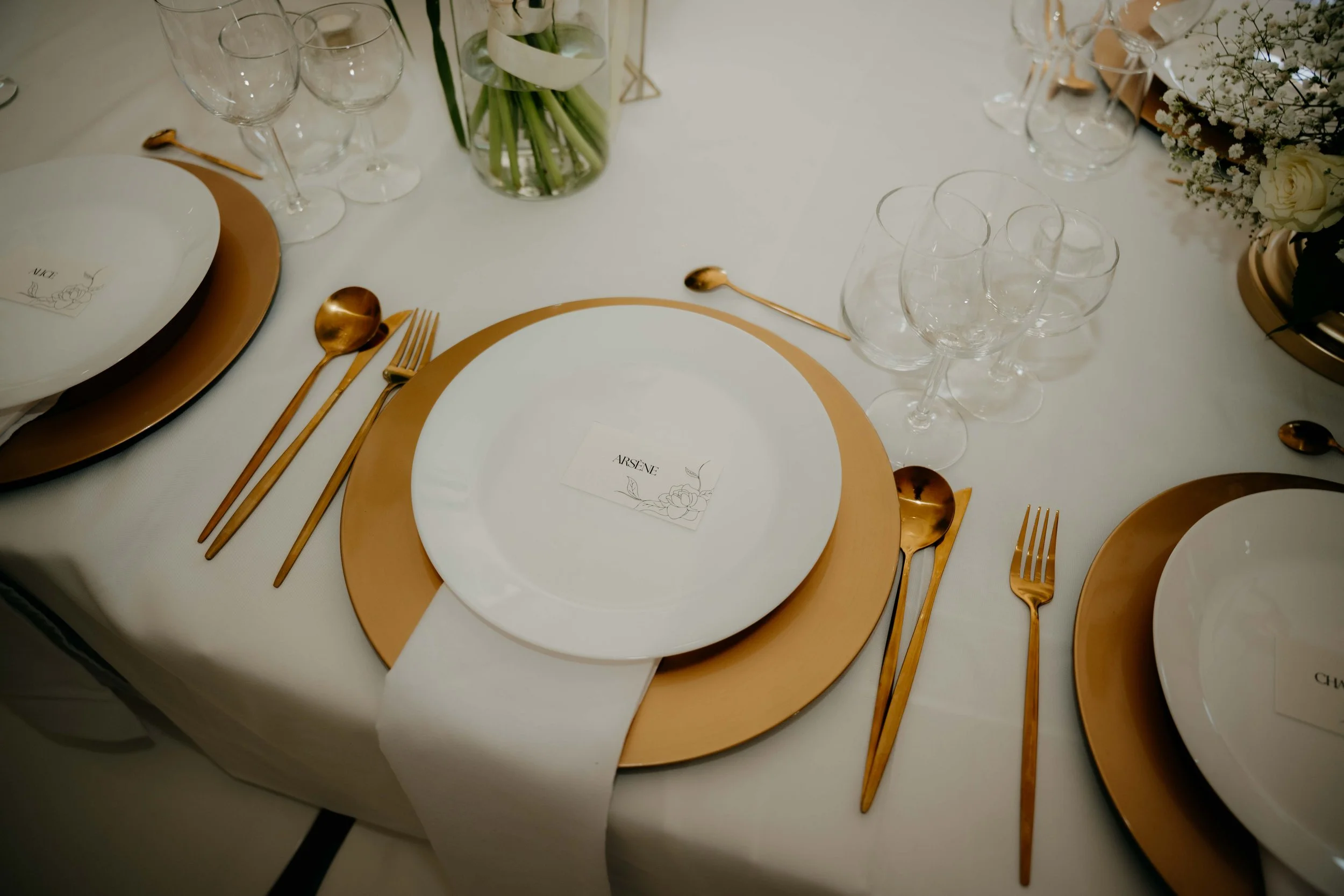 A formal table setting featuring white plates with gold chargers, gold utensils, and clear wine glasses. In the center, there is a small arrangement of flowers including white roses and baby's breath, with green stems in a glass vase. A place card wi