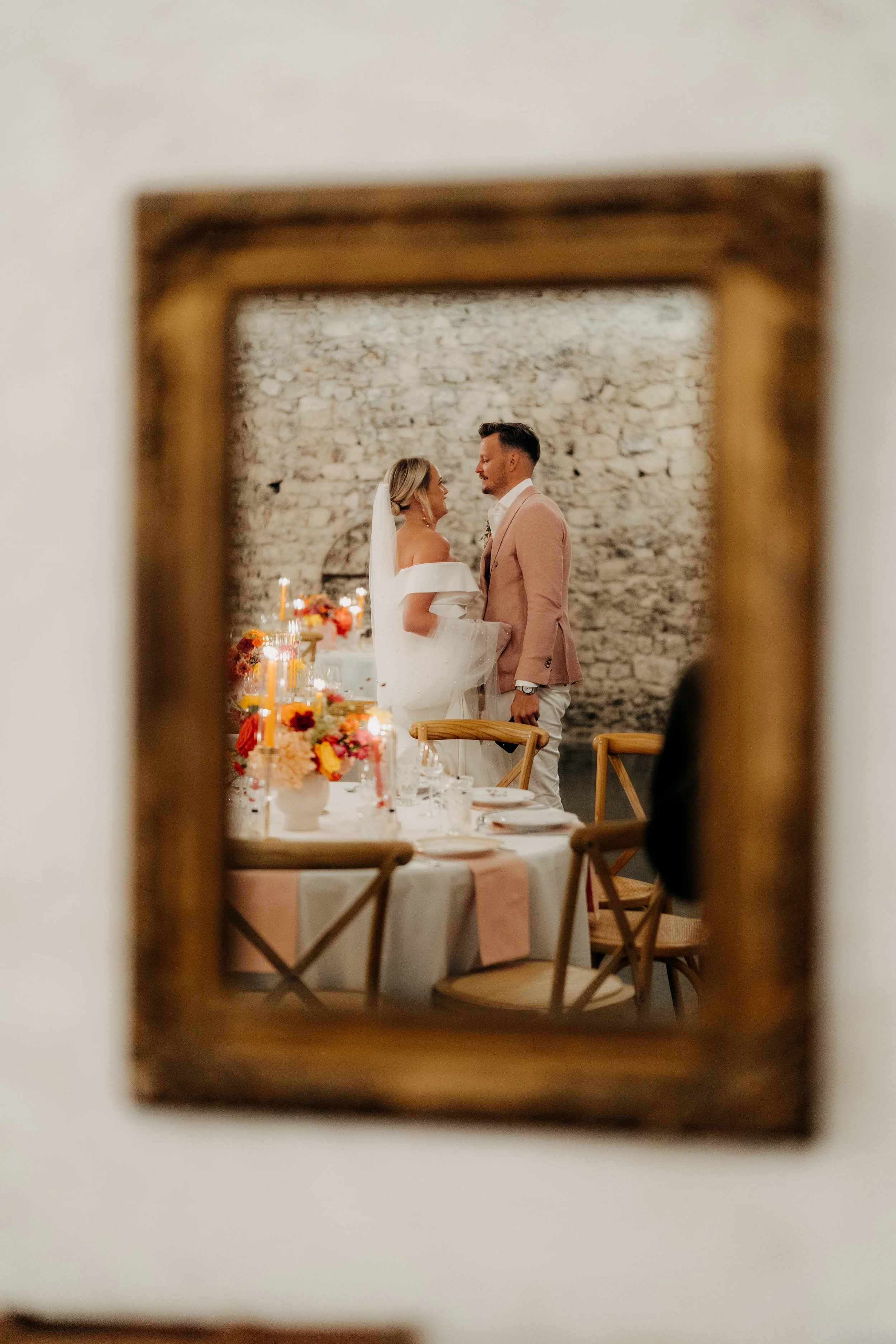 Reflection of a bride and groom standing facing each other during a wedding reception, seen through a small wooden framed mirror. The background features a textured stone wall and a table decorated with colorful flower arrangements.