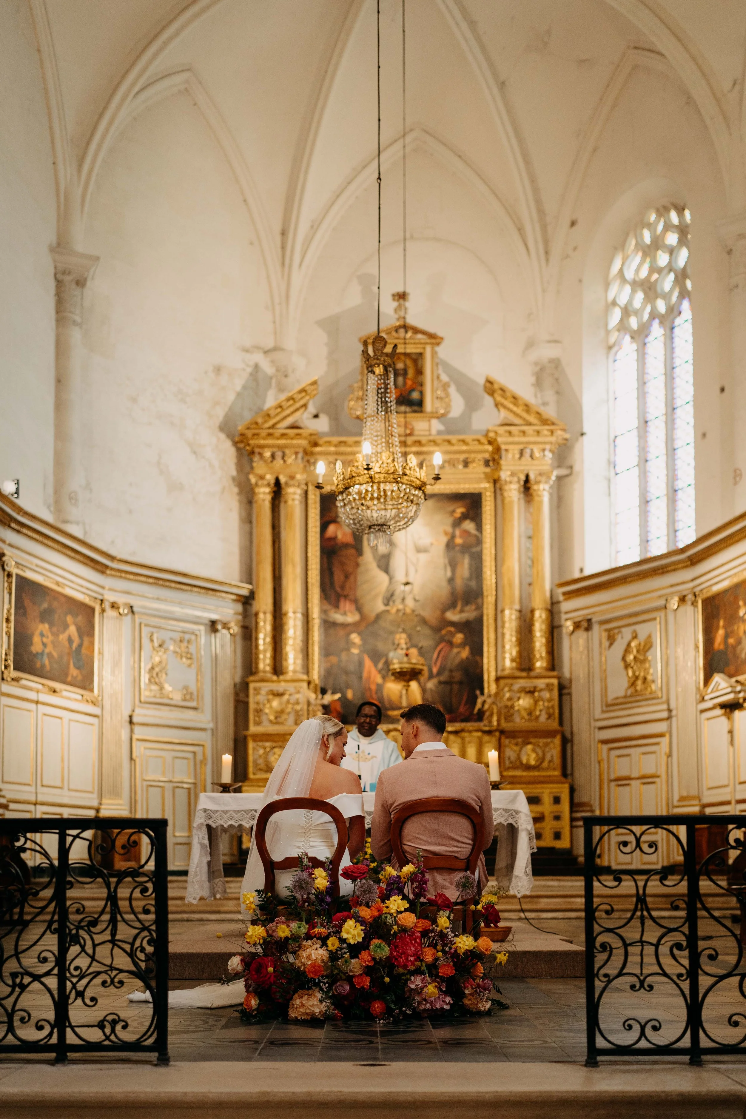 A wedding ceremony inside a church with ornate gold and white decorations, stained glass windows, and a large chandelier. The bride and groom are seated at an altar, facing a priest. The altar is decorated with a large colorful floral arrangement at 