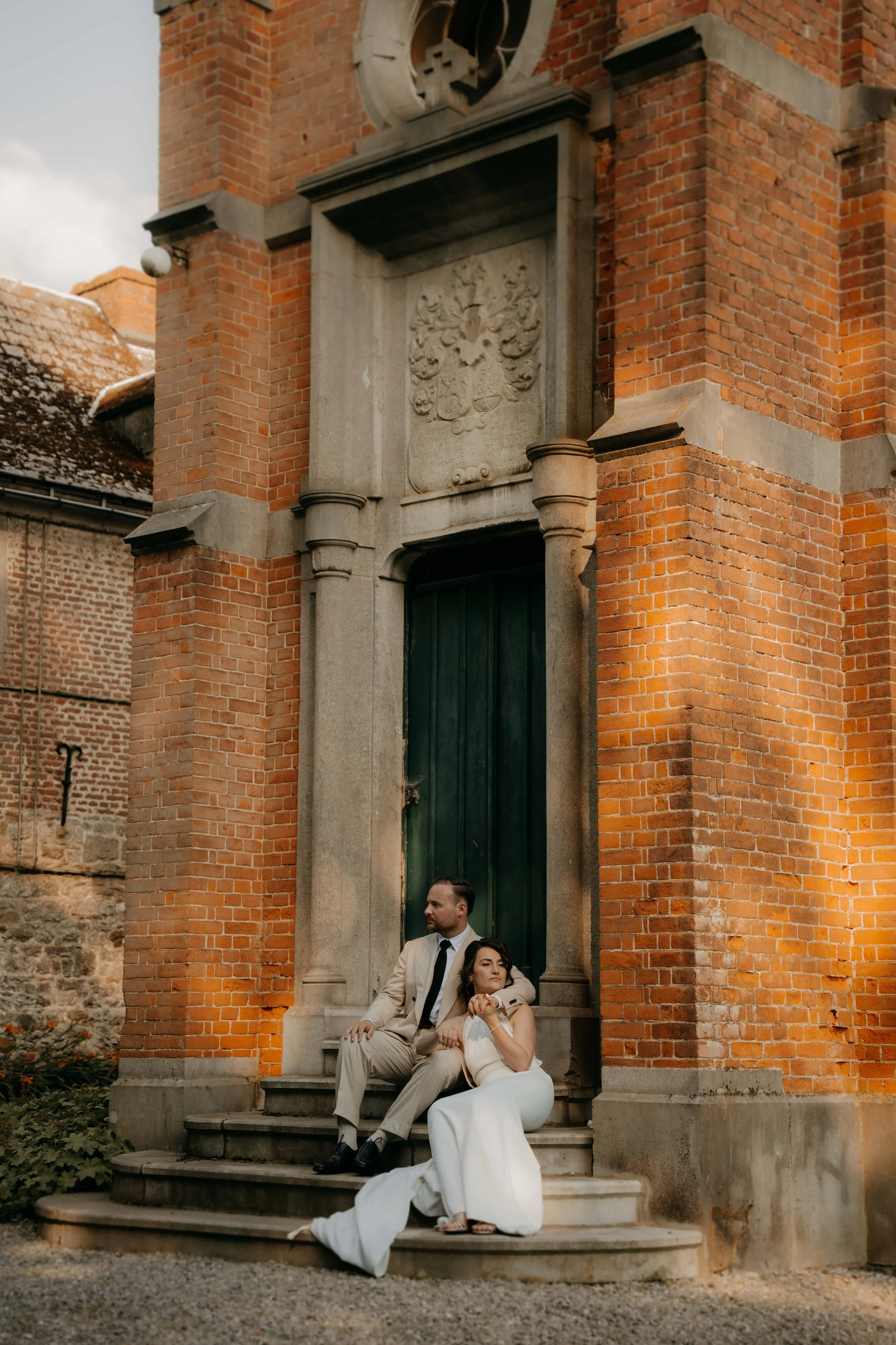 A man and woman in formal attire sitting on the stairs outside a brick building with a crest above the door. The man is dressed in a beige suit with a black tie, and the woman is in a white dress. They are seated next to each other, with the man lean