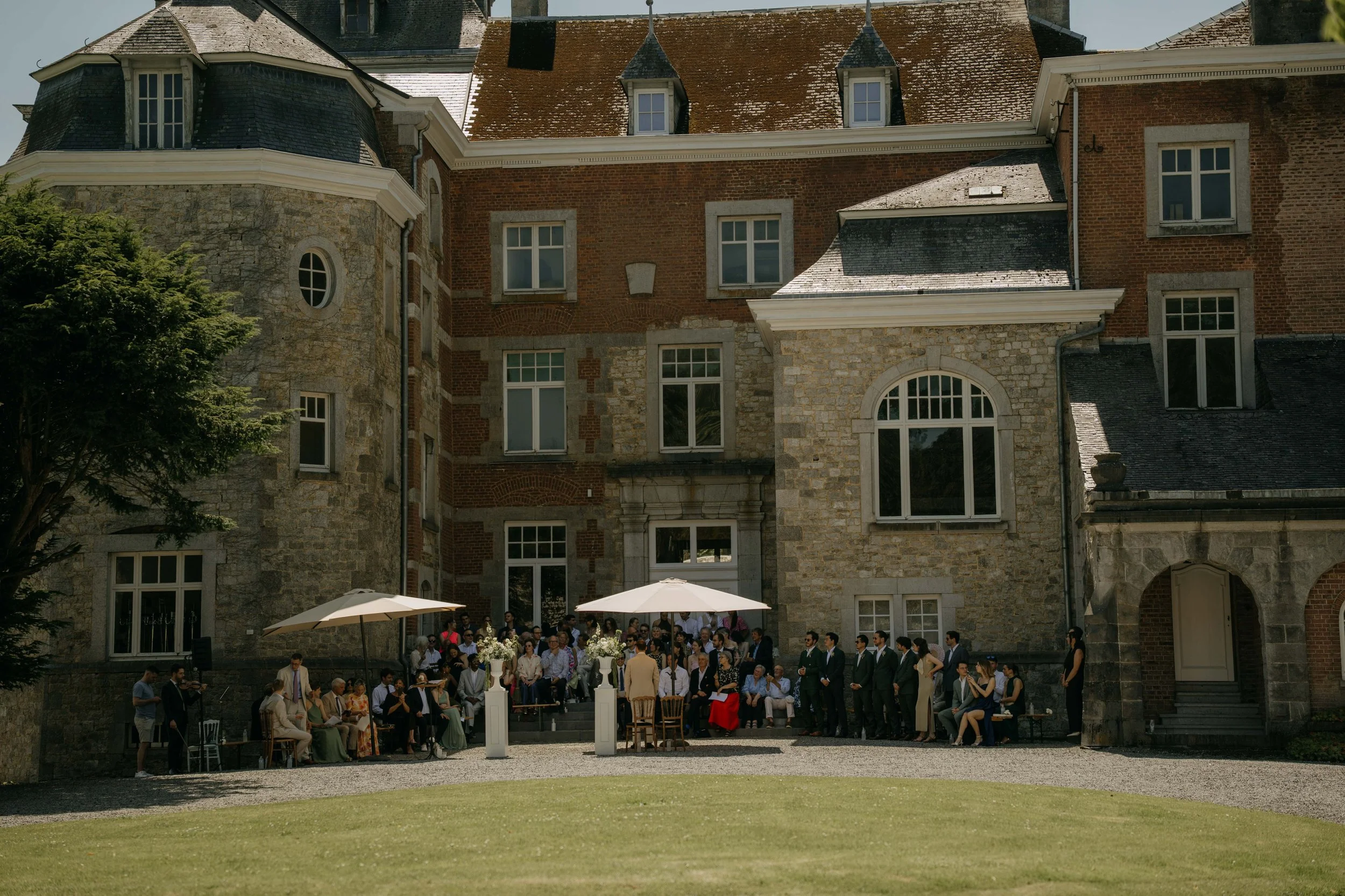 A large group of people gathered outside a historic brick and stone building during daytime, with some sitting and others standing under umbrellas, possibly attending a social event or wedding.