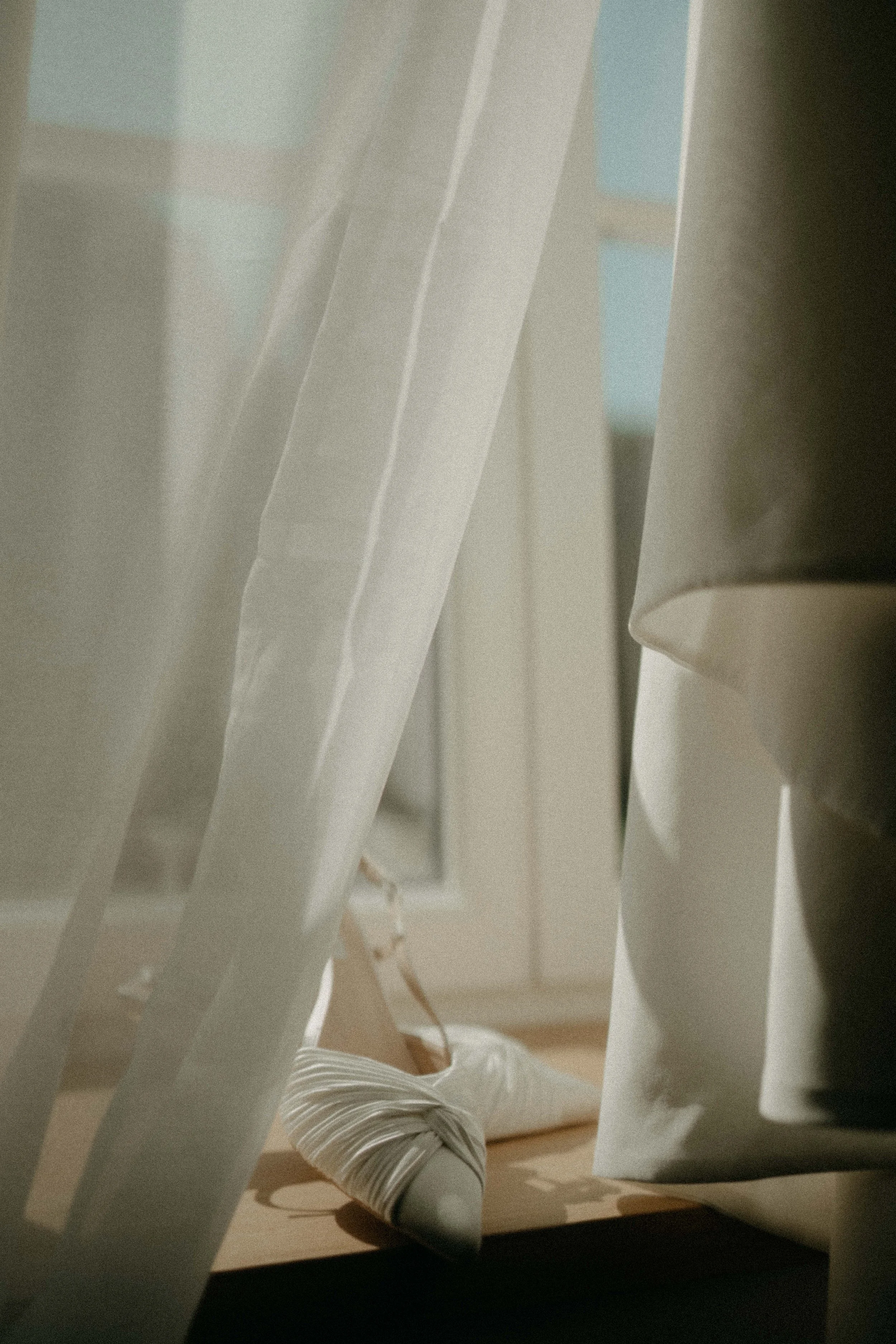 A person lying on the floor with legs crossed, wearing white and gray striped pants and white shoes, viewed through white vertical blinds with sunlight filtering through.