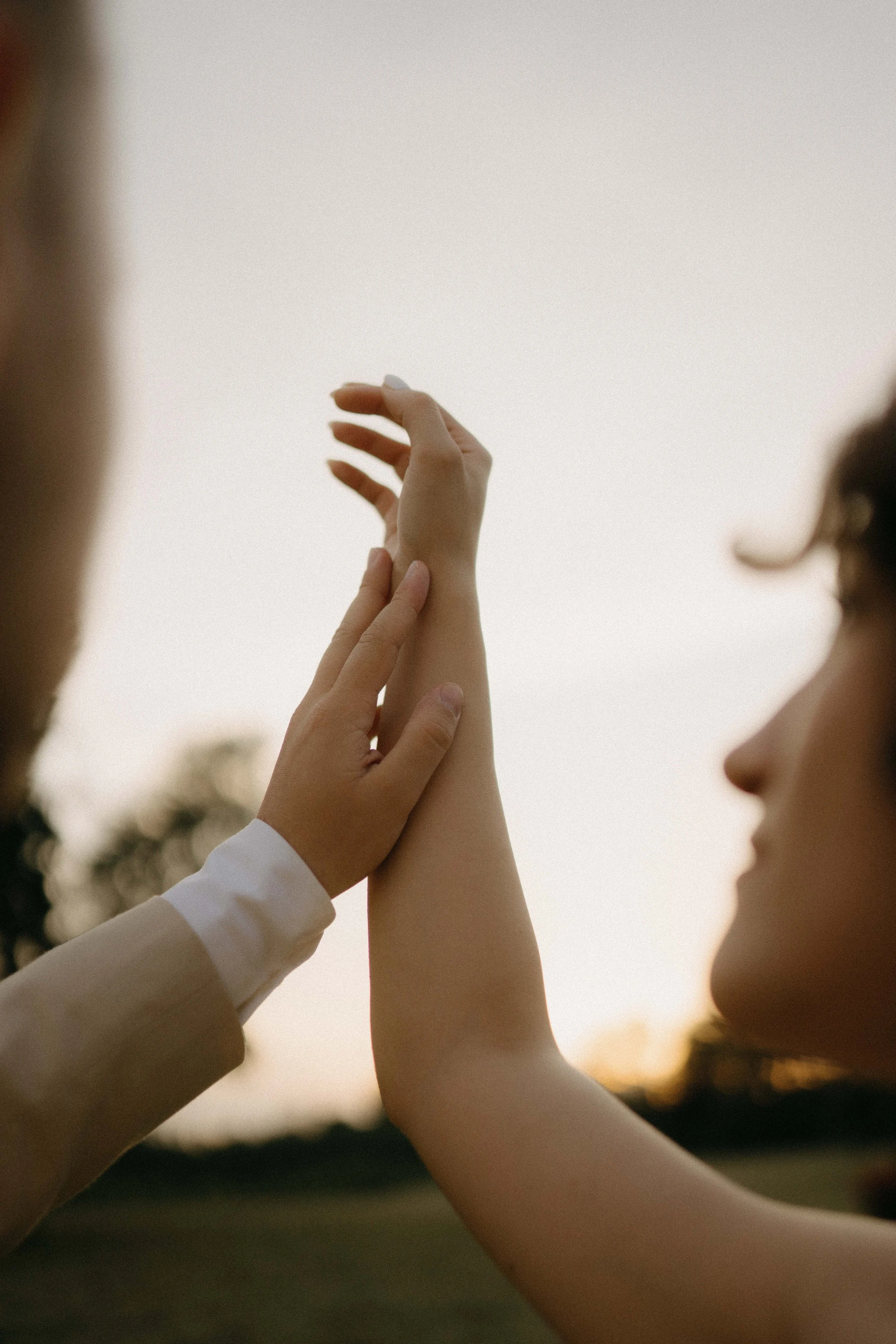 Two women holding hands and touching fingers, with a blurred background and sunset sky.