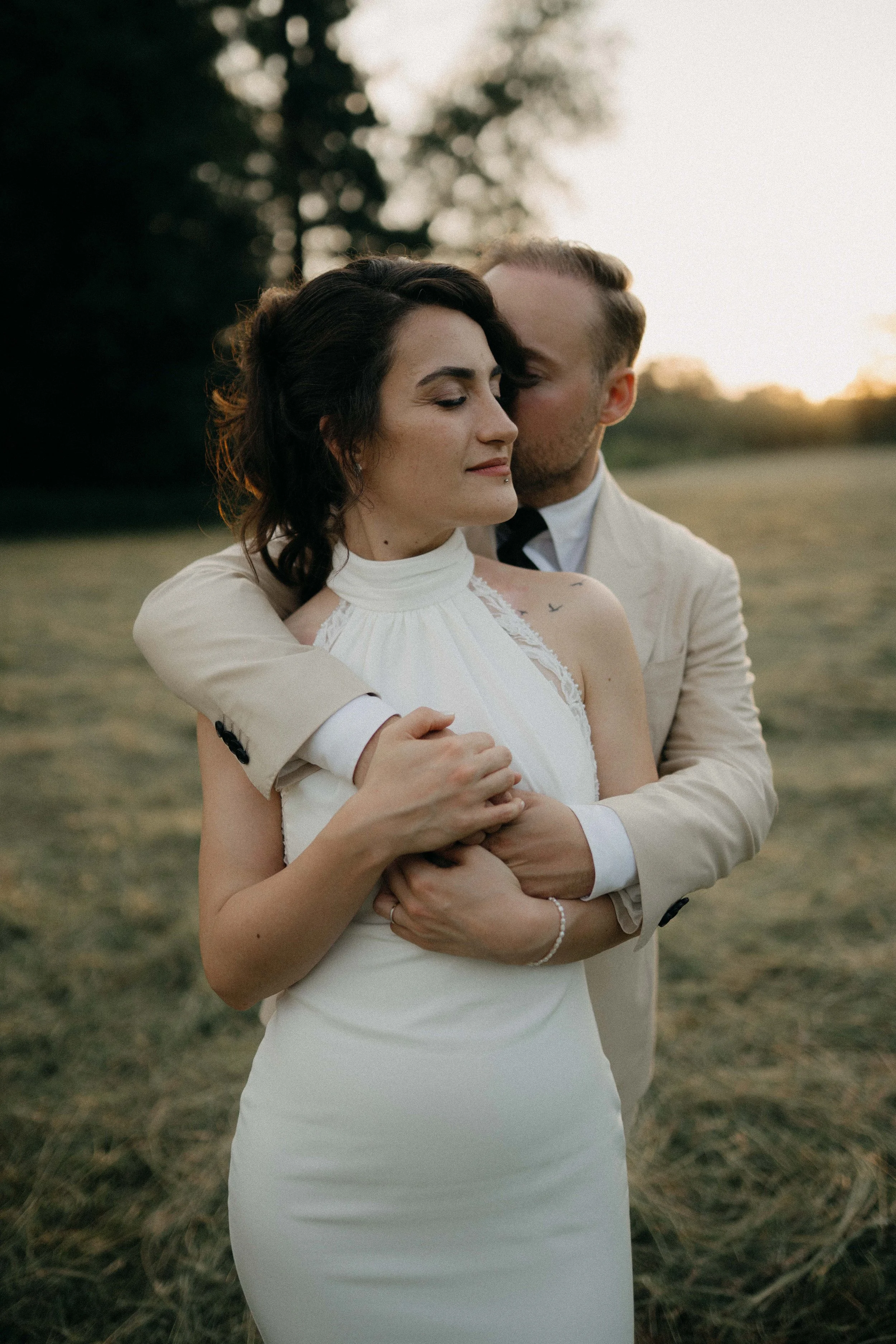 A couple in formal attire embracing outdoors at sunset, with trees in the background.