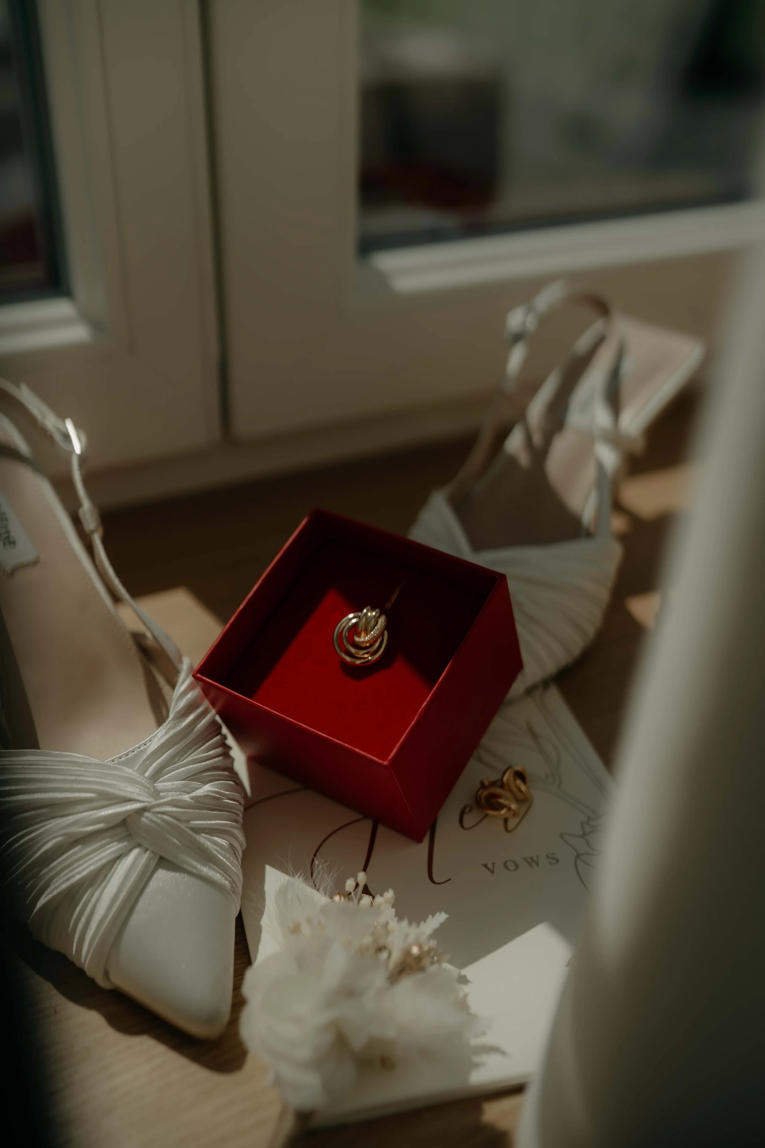 Close-up of jewelry and accessories, including a red box with rings, white shoes with ribbon decoration, and flowers, on a wooden surface.