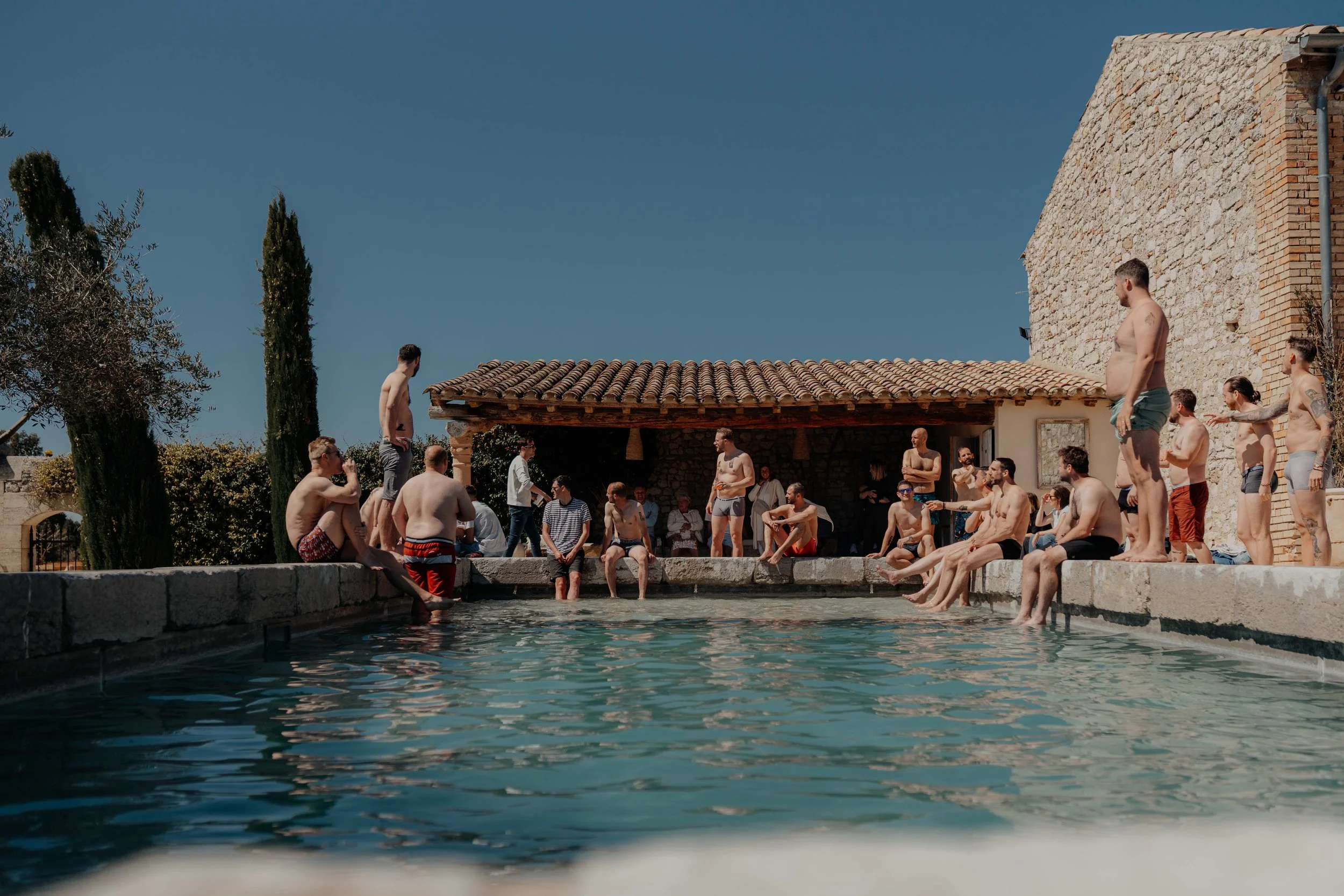 People relaxing by a pool, some sitting on the edge and others standing, with a rustic building and tall trees in the background under a clear blue sky.