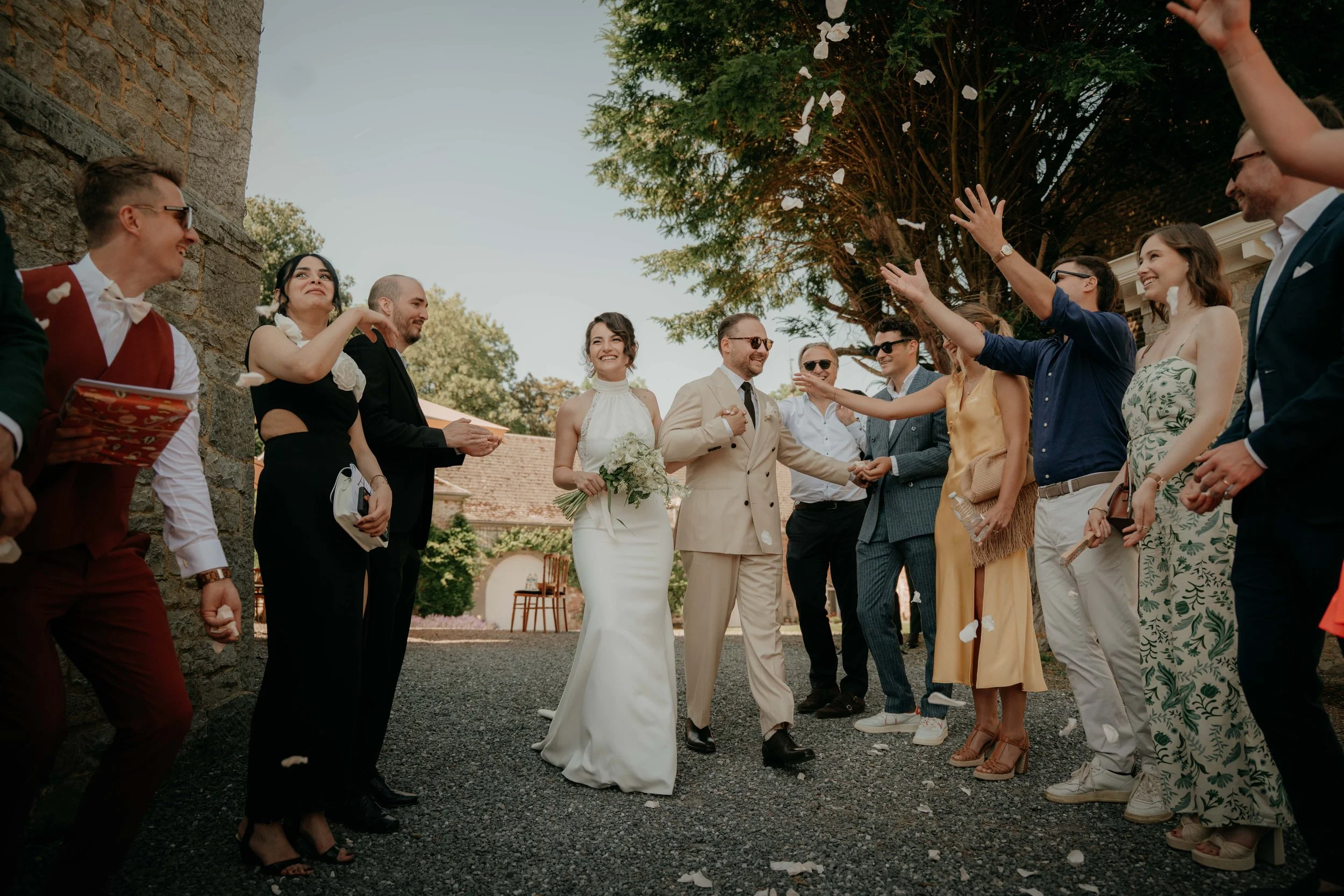 A bride and groom at their wedding reception outdoors, celebrating with friends and family under a tree with white flowers, with guests throwing flower petals.